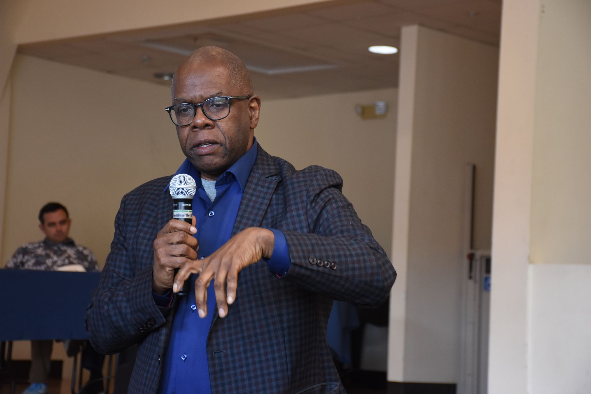 Man speaking into microphone, gesturing with his hand. He is wearing glasses, a blue shirt, and a dark jacket.