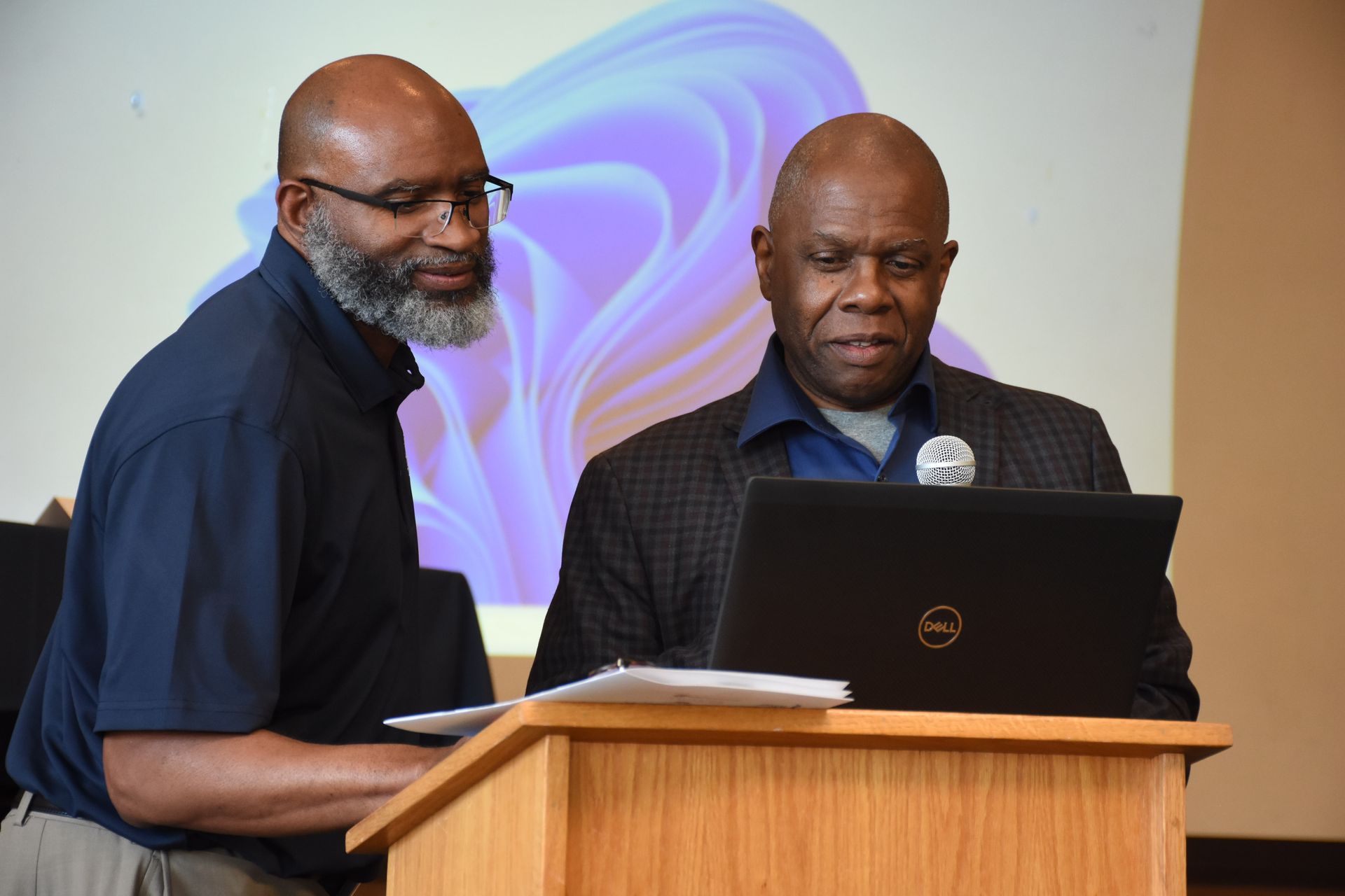 Two men beside a podium with a laptop; one looks at the screen. A Windows 11 background is visible.