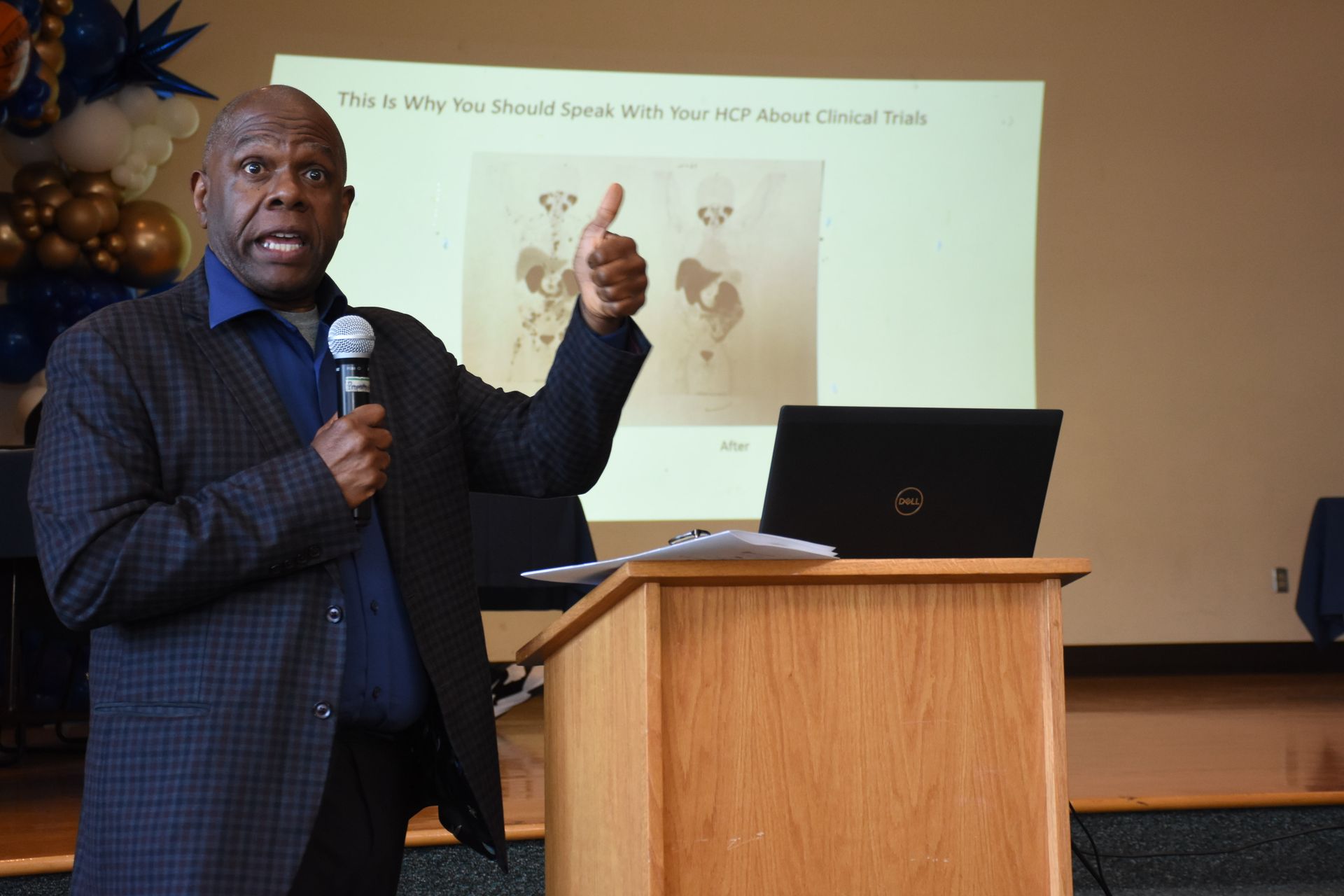 Man presenting with microphone, pointing at a screen showing medical images. He's standing behind a podium in a room.