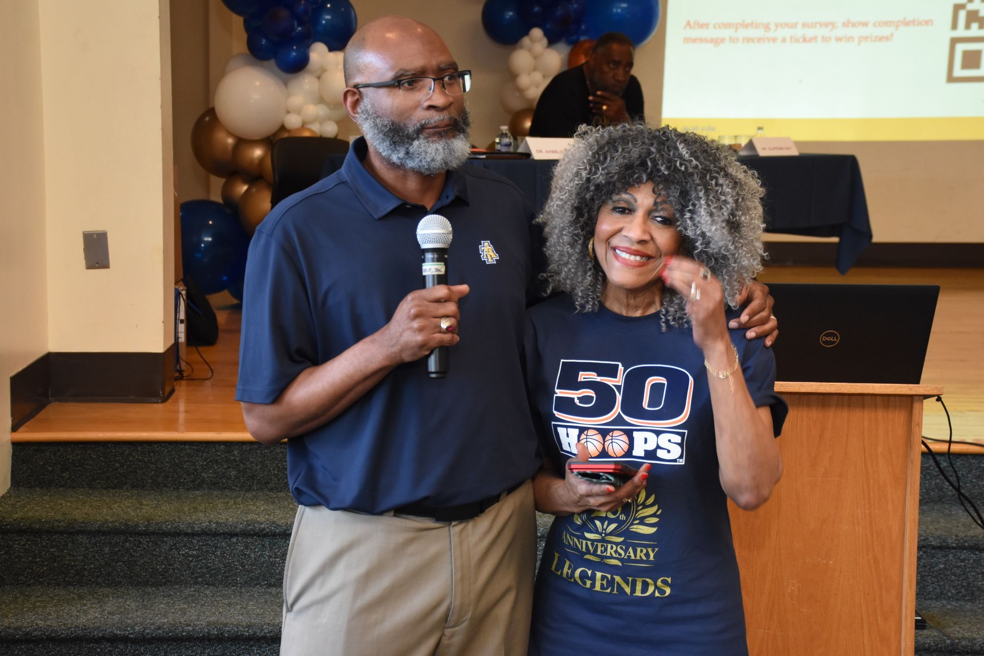 Man and woman on a stage; man holds a microphone, woman has arm around him, both smiling. Background has balloons.