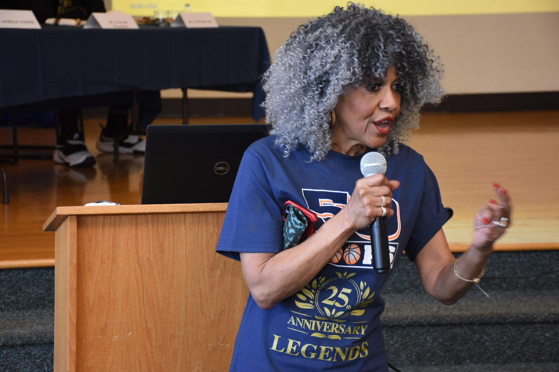 Woman speaking into a microphone, standing behind a podium. Wearing a navy blue shirt with a graphic, gray hair.