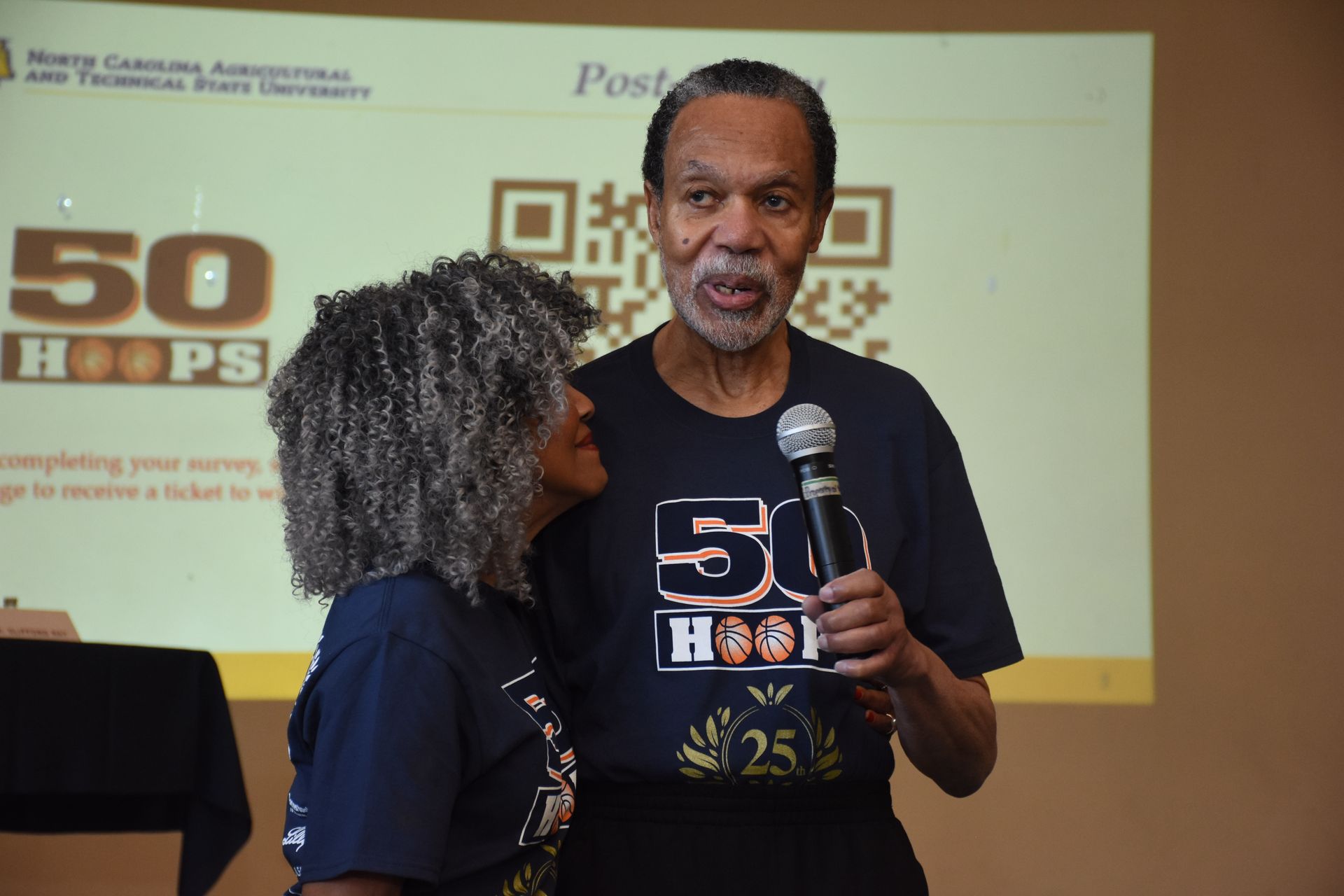 Two people, a man with a microphone and a woman, stand together in front of a presentation screen, promoting 