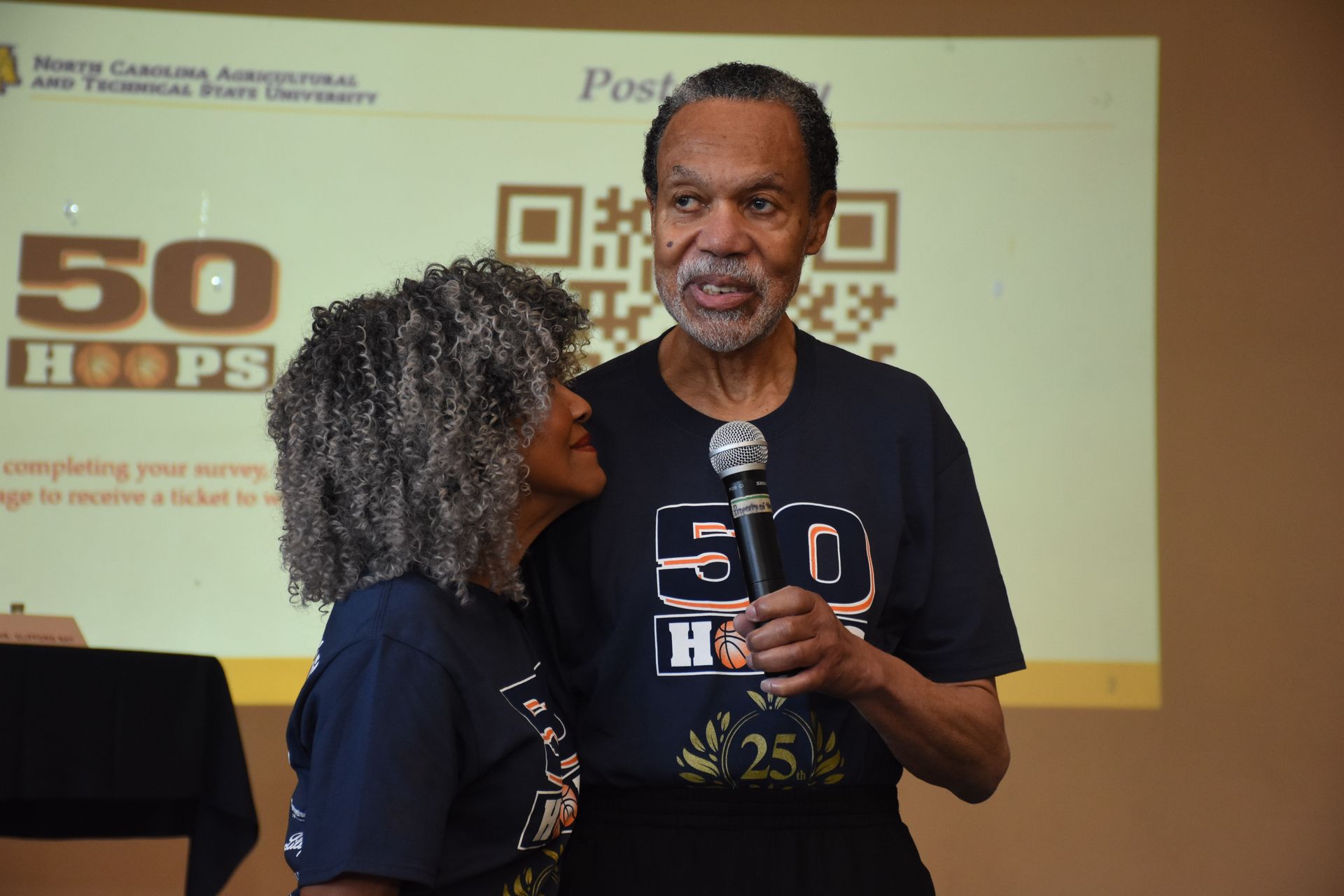 Two people standing near a screen with text, both wearing navy blue shirts with 