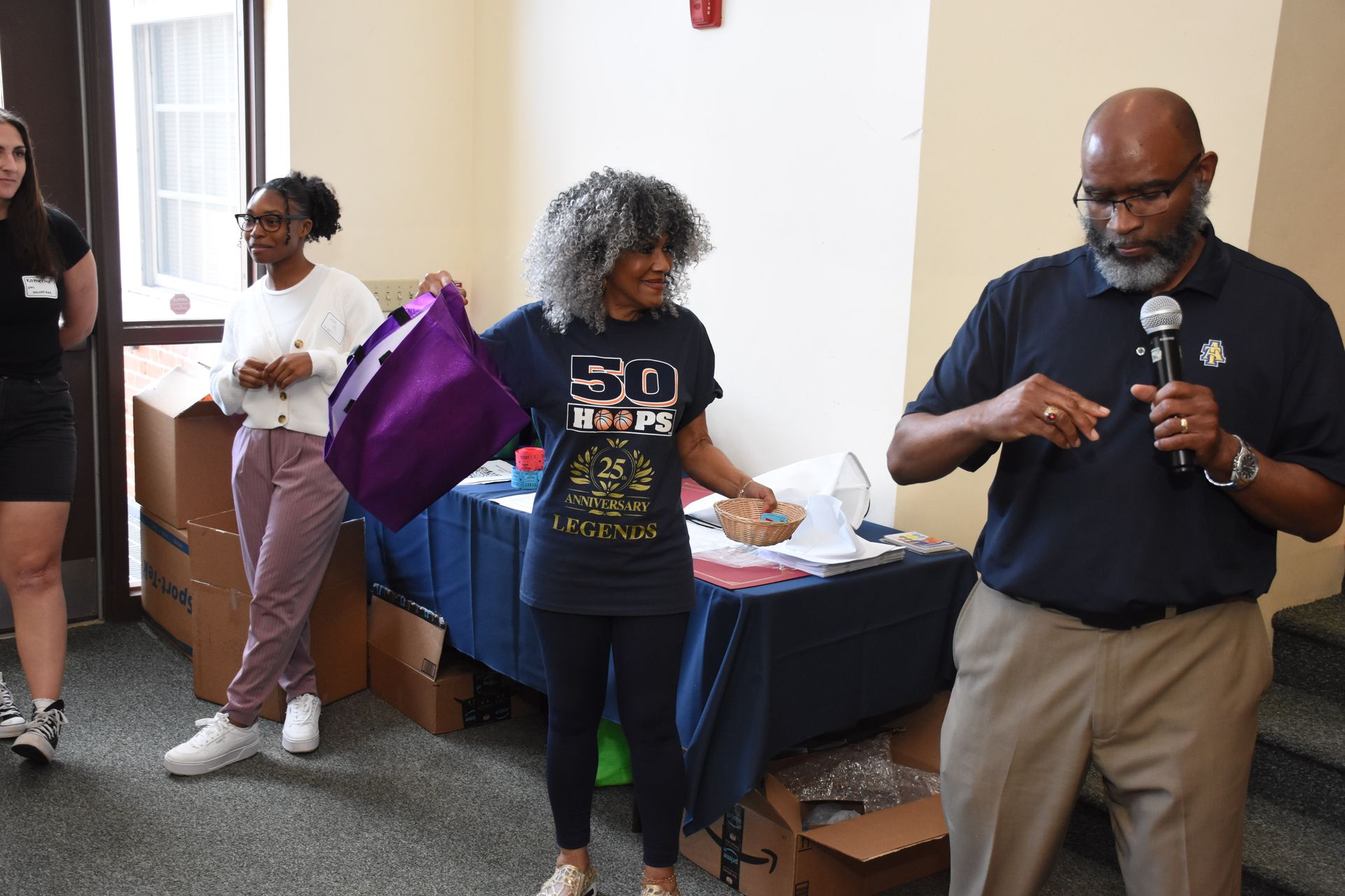 Man speaking into a microphone. Woman holds purple bag. Two people stand behind a table, boxes in the background.