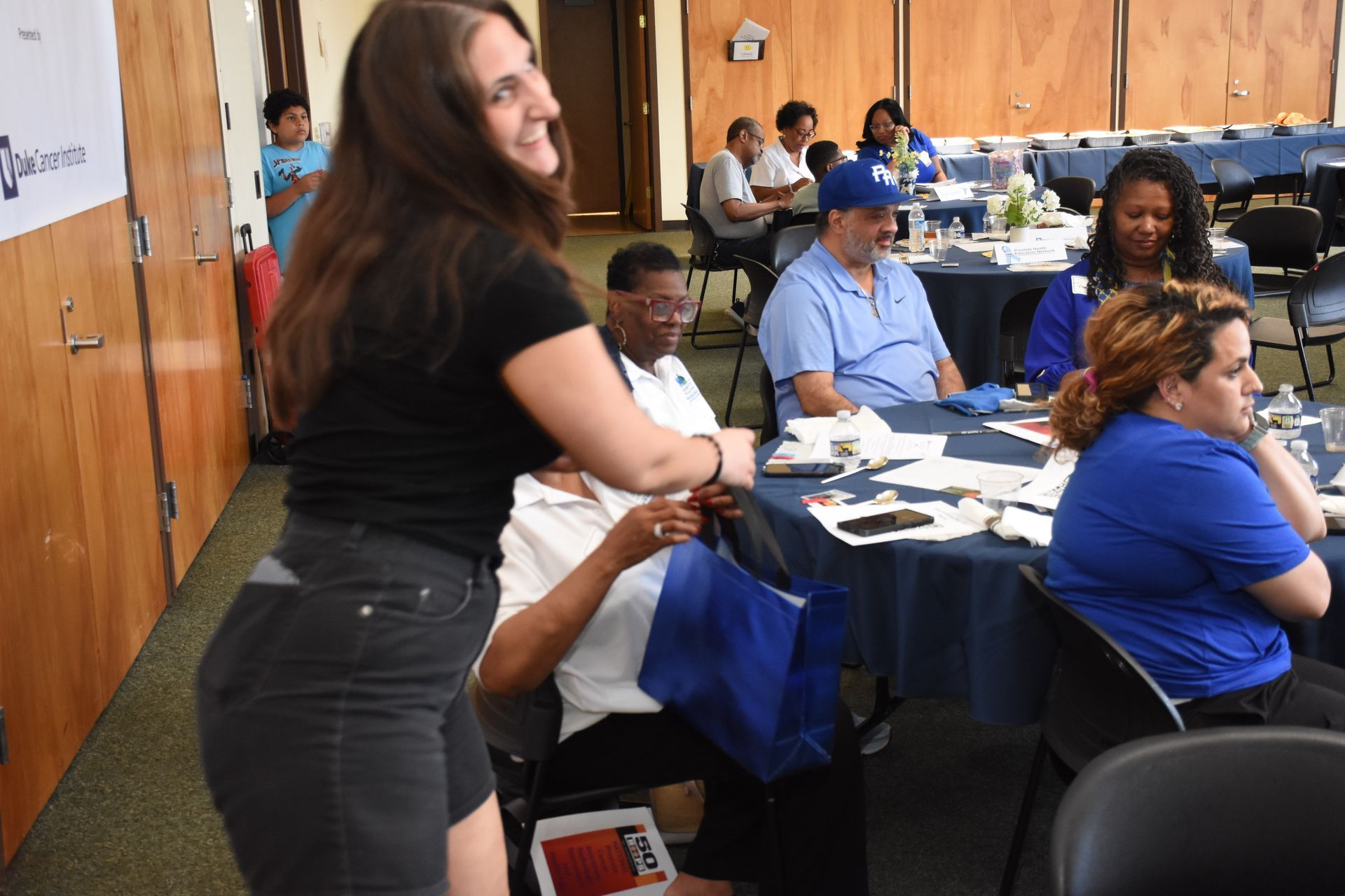 Woman smiling, holding a blue bag, walking through a room with people seated at tables, some looking at her.