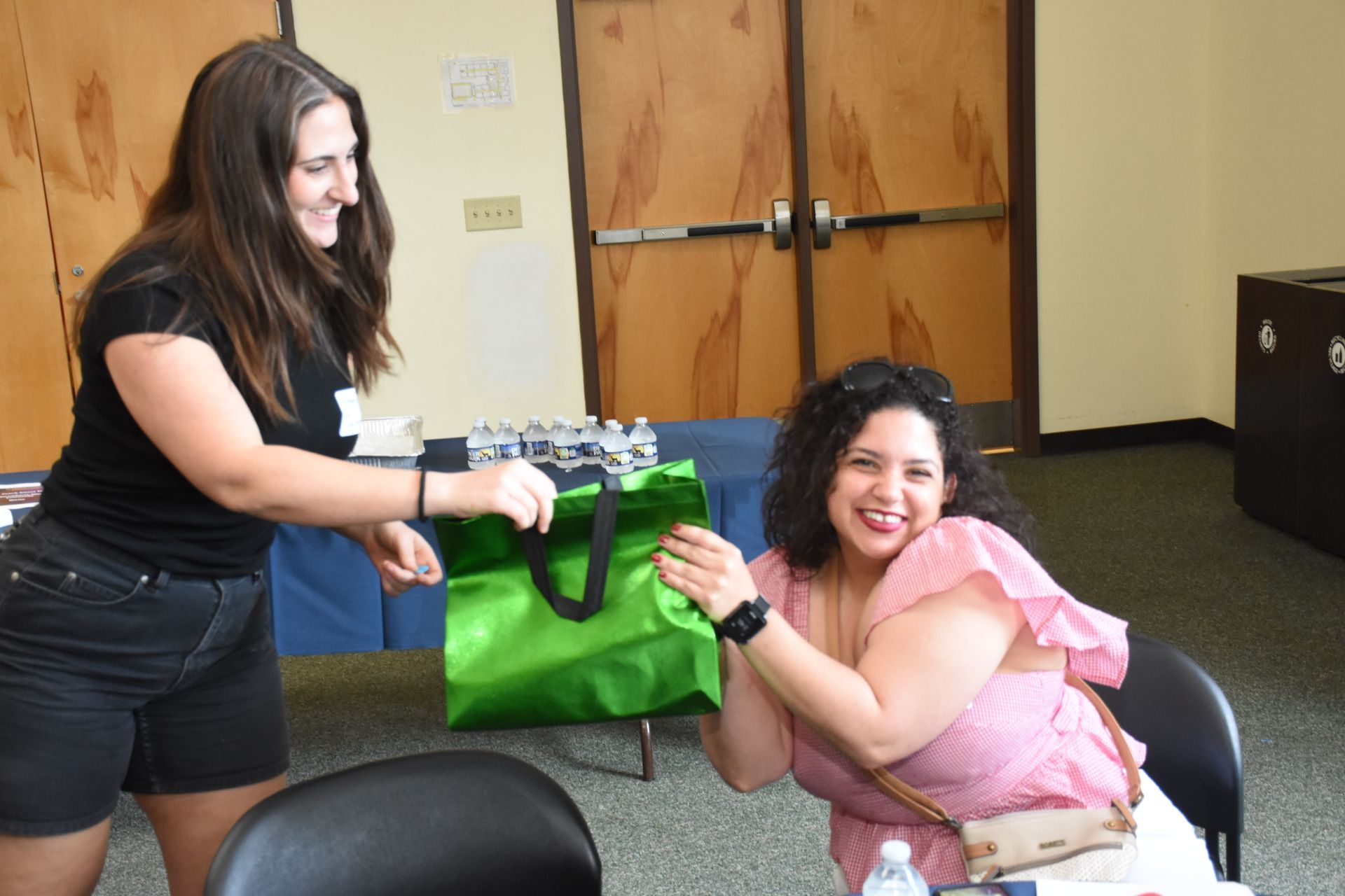 Woman handing a green bag to a seated, smiling woman. Both are indoors near a table.