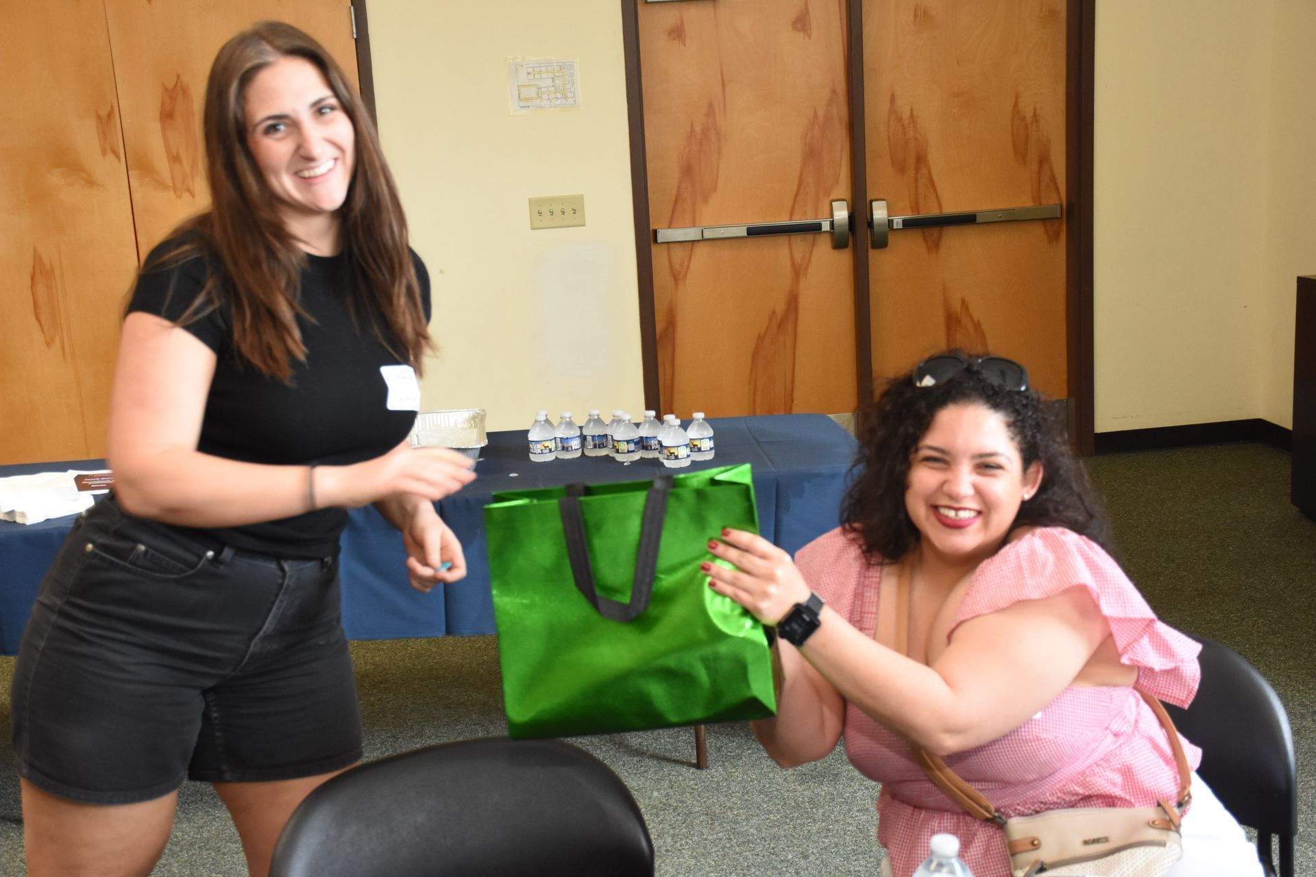 Two women smile, one hands a small item, the other holds a green bag. Inside a room with wood paneling.