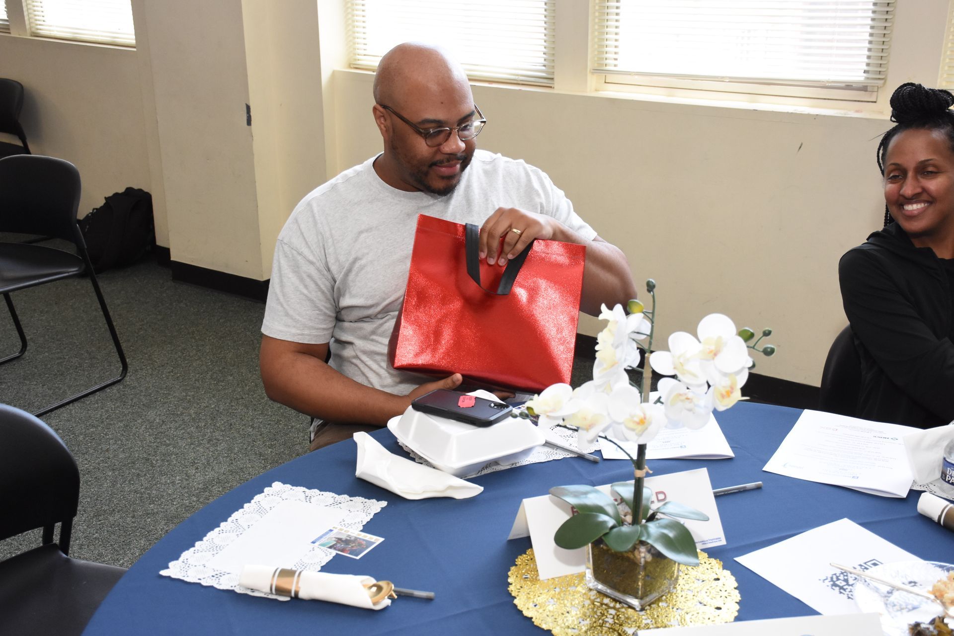 Man opening a red gift bag at a table with flowers, smiling woman at his side.