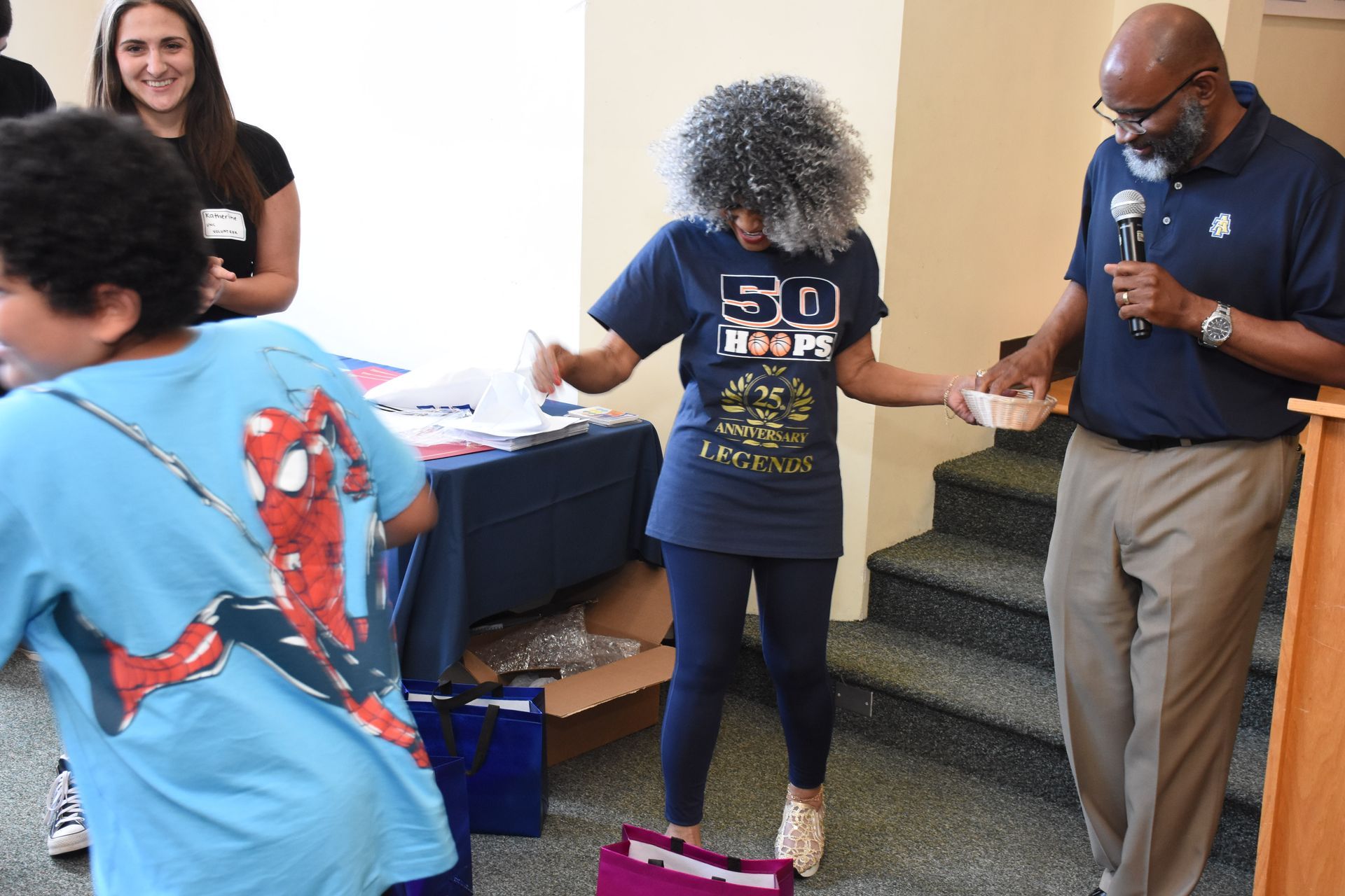 A man with a microphone hands a prize to a woman at a gathering. A child in a Spiderman shirt runs by.