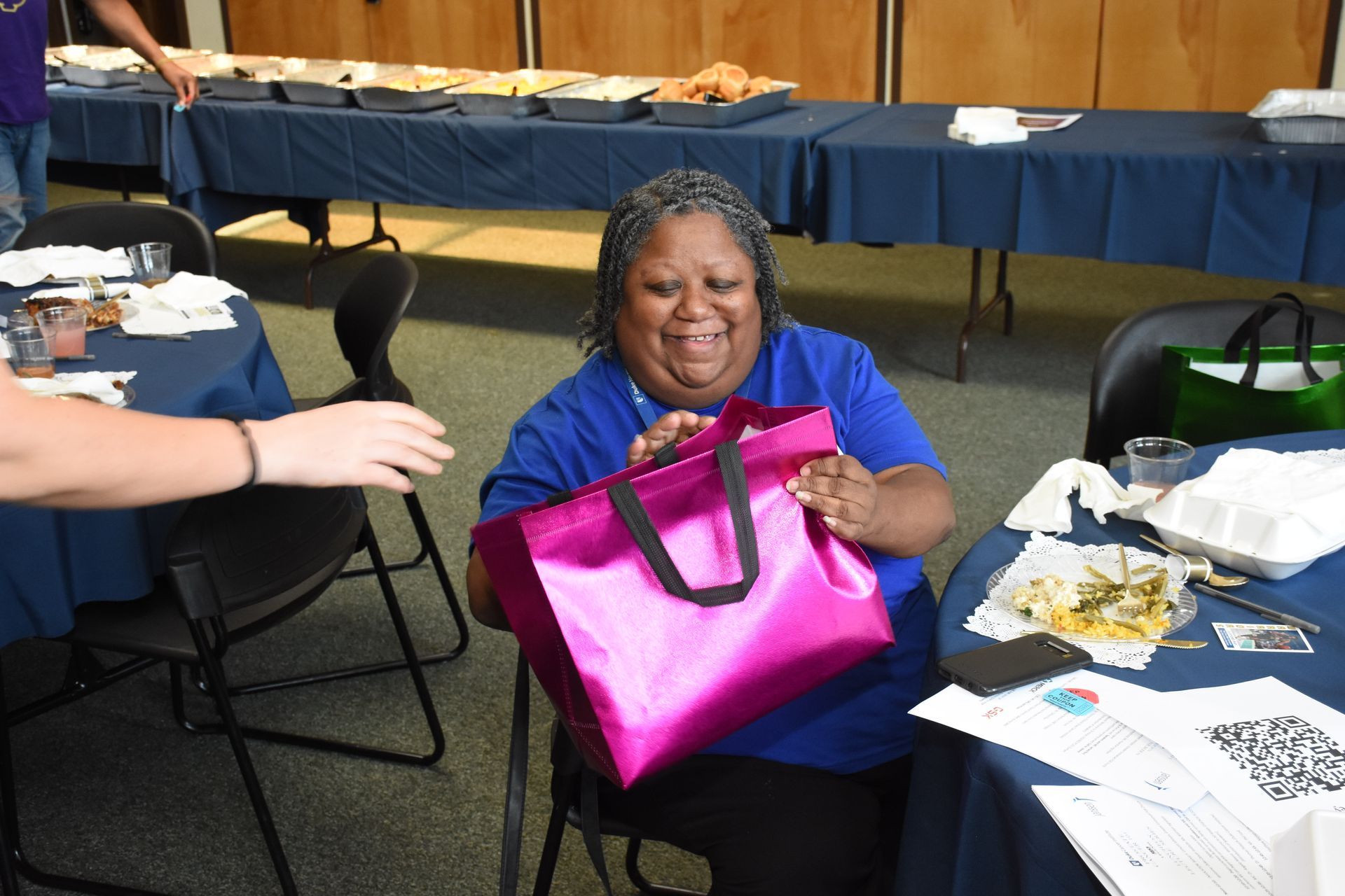 Woman smiles, holding a bright pink bag. A person hands her the bag at a table with food.