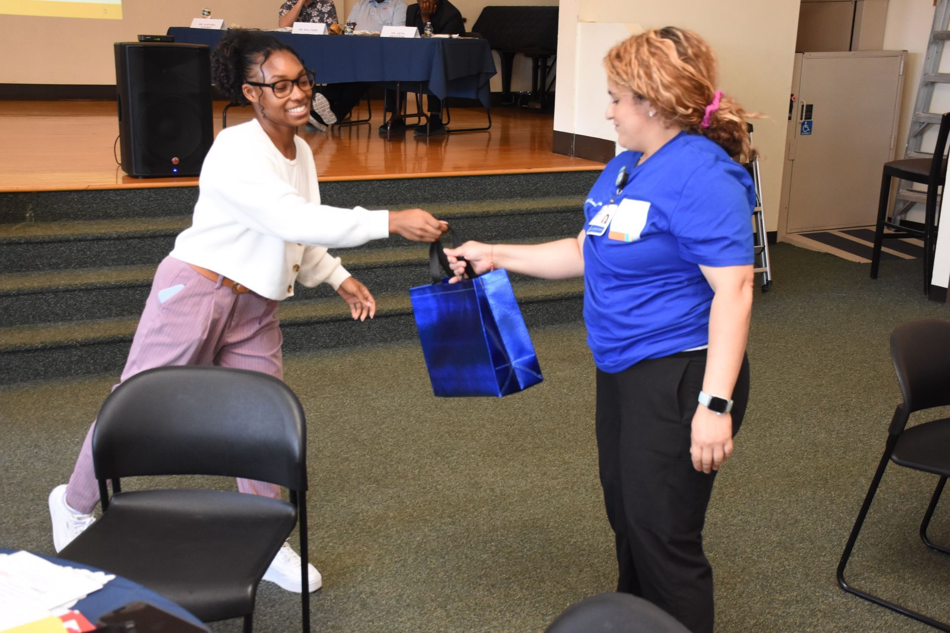 Person in blue shirt handing gift bag to person in white shirt. Inside a room with chairs and stage.