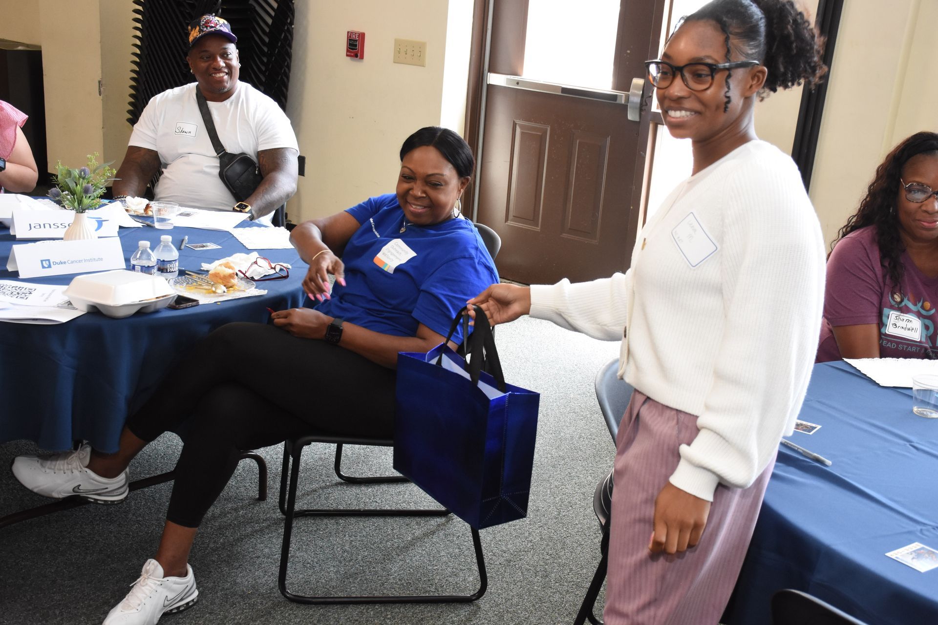 Woman hands a blue tote bag to another woman at a table in a meeting. Other people are in the room.