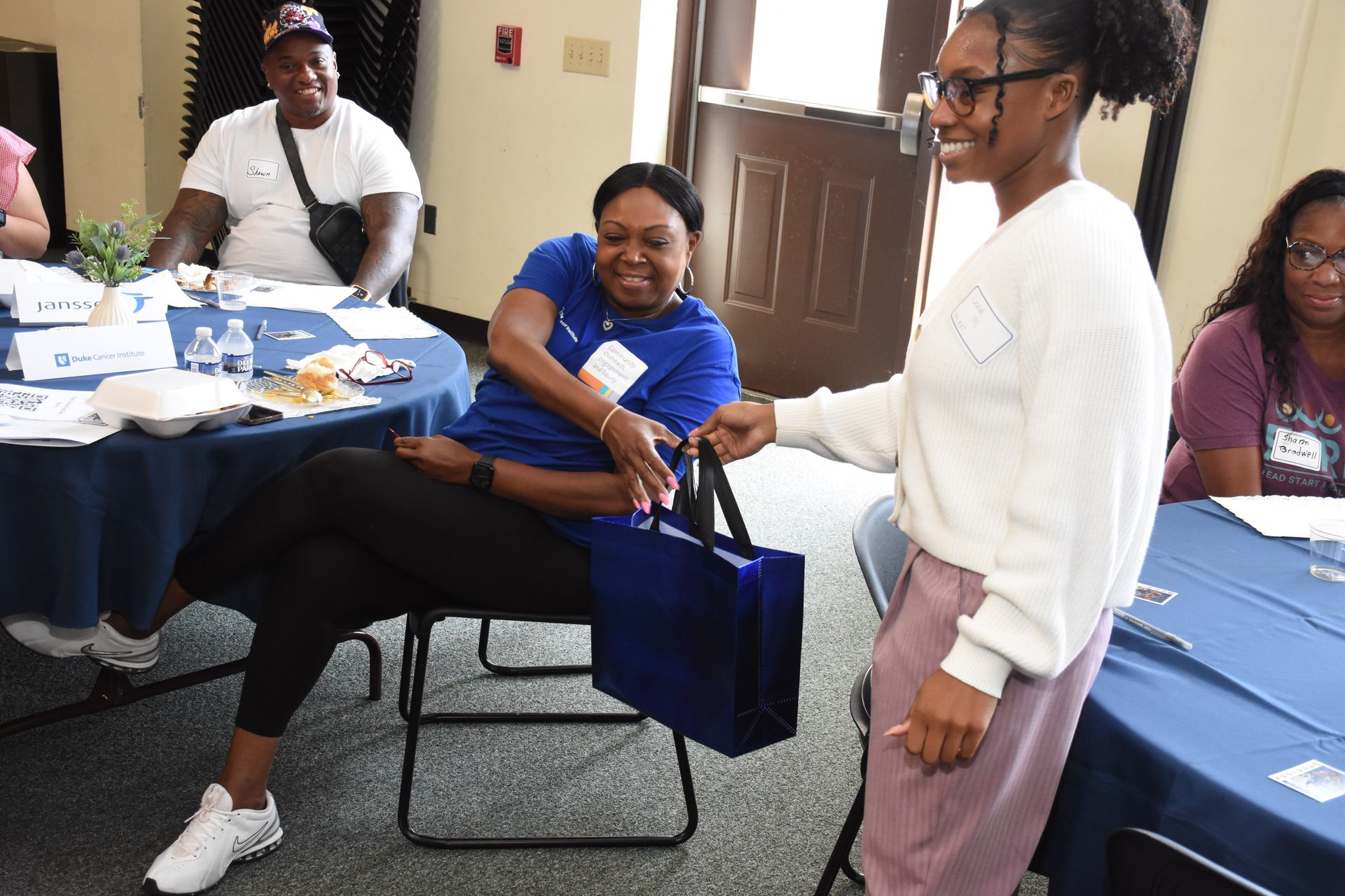 Woman seated at table receives a blue bag from another woman in a meeting setting.