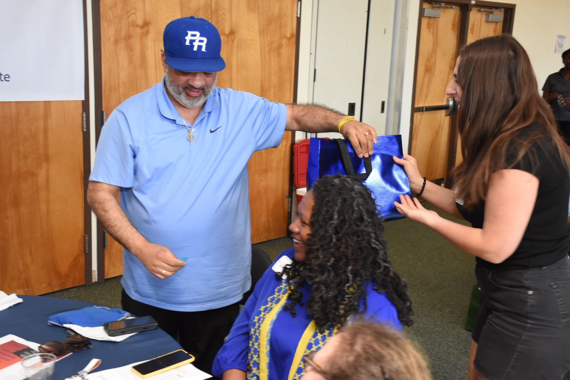 Man in blue cap/shirt gives gift bag to a woman in a blue dress, assisted by another woman. Indoors.