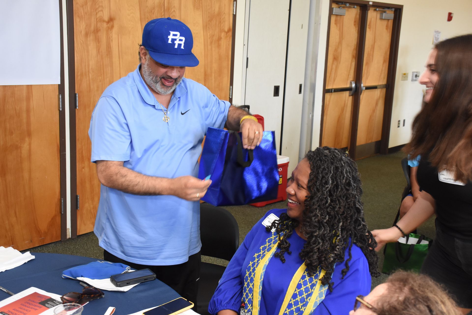 Man in blue shirt and hat shows items to smiling woman, others watch. Indoors, near table.