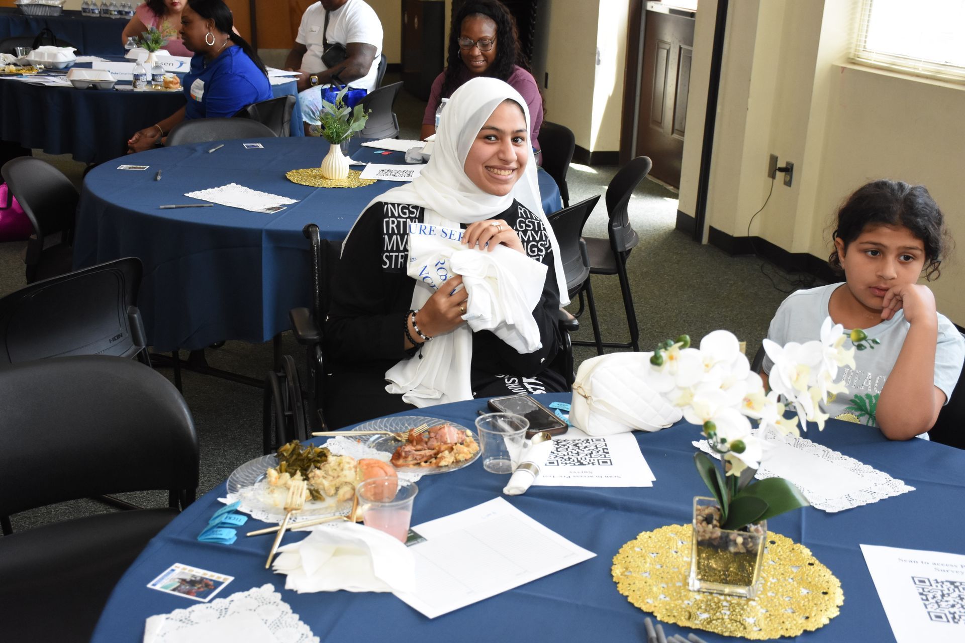 Woman at table smiles, holding napkins; child looks on. Event with food, blue tablecloths, other attendees.