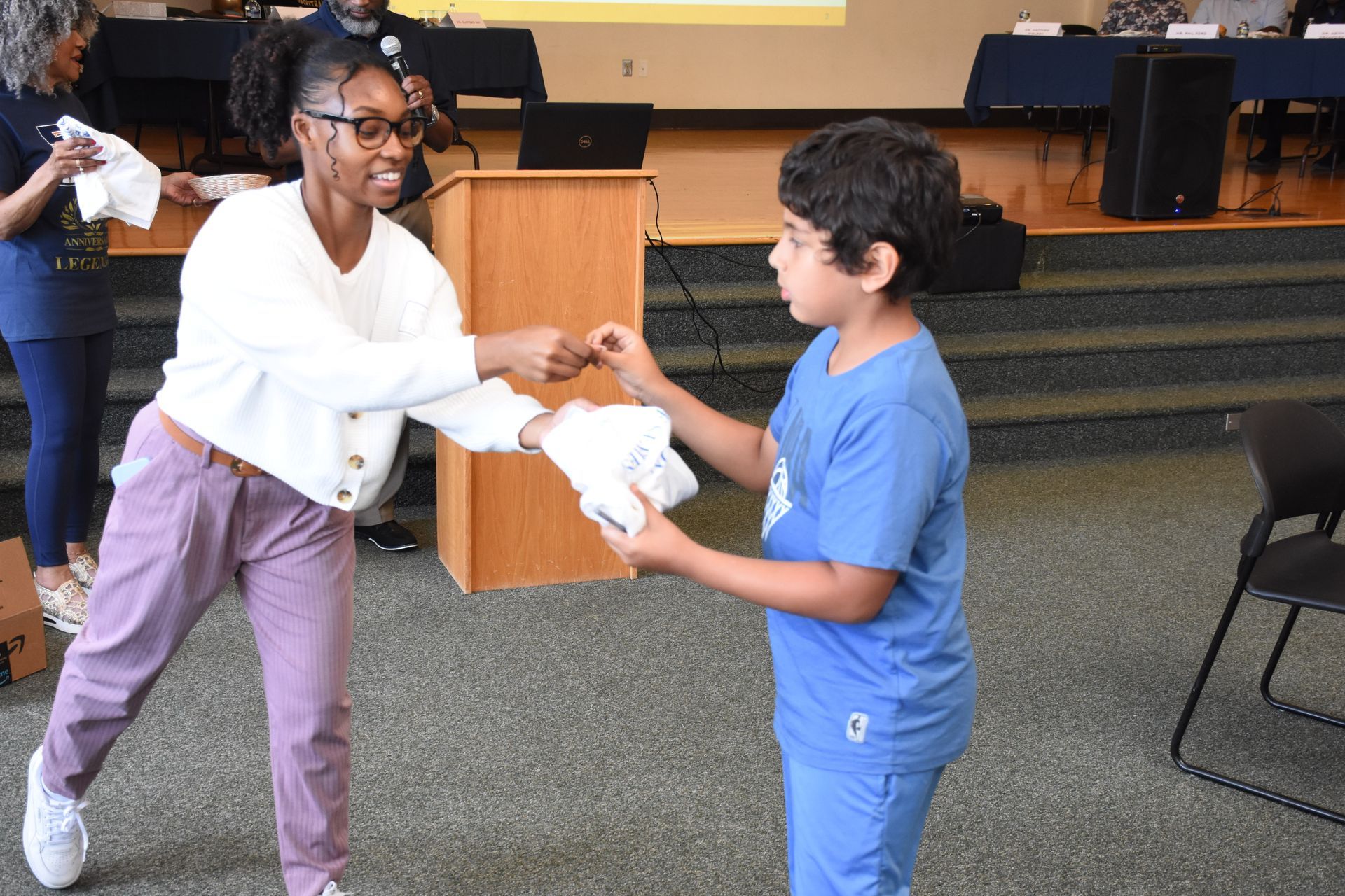 A woman in white and purple pants handing a child in blue a white item. They are in a room with a podium.