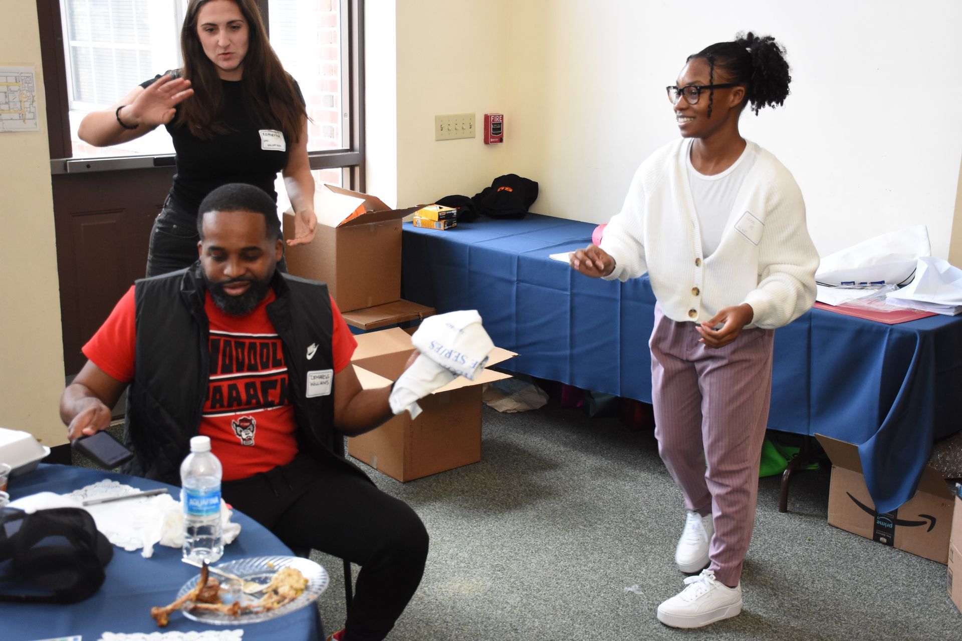 Three people in a room: a person seated at a table, a person standing and smiling, and another person reaching back.