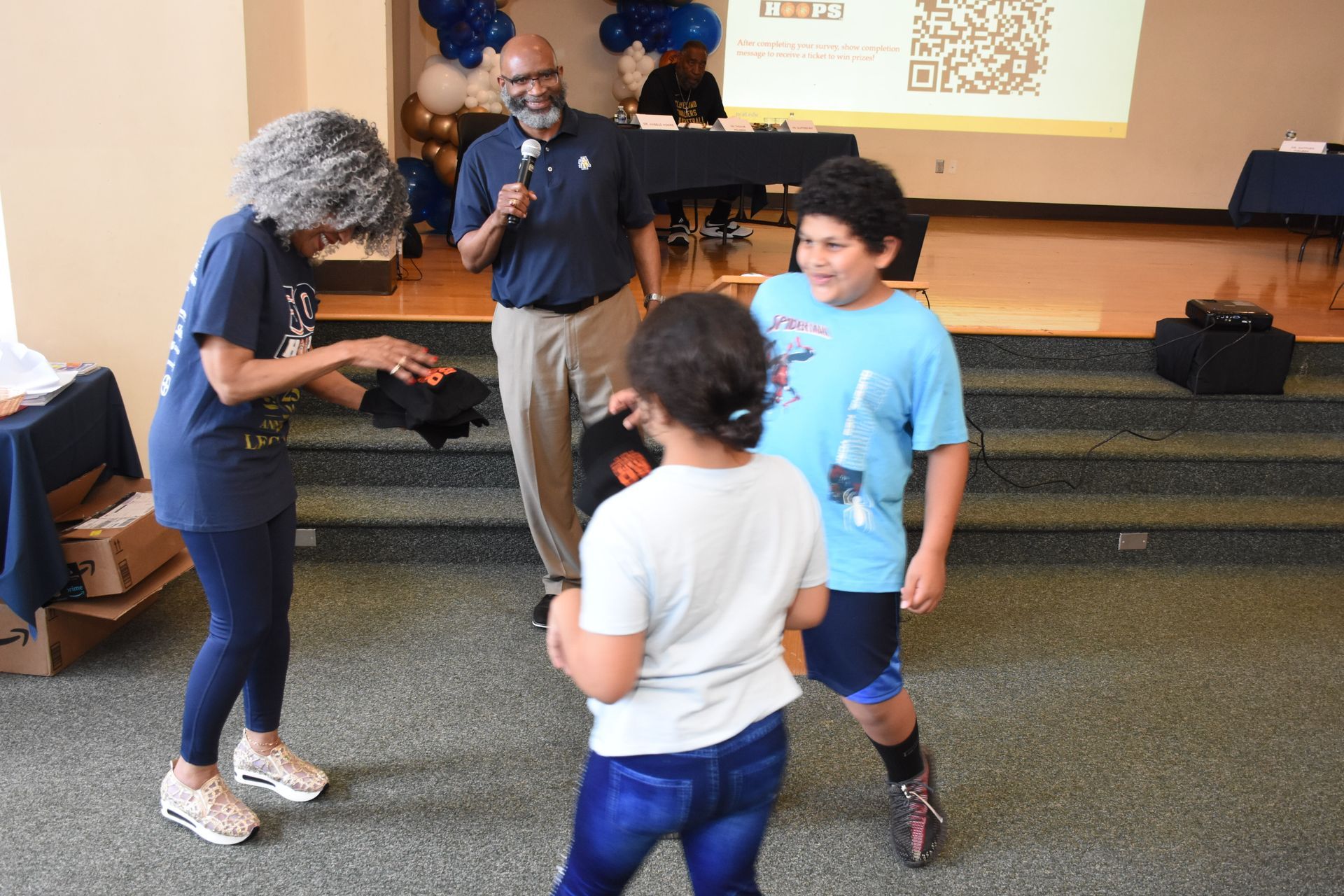 Woman hands out items to children at an event. Man speaks into a microphone. Room with a stage.