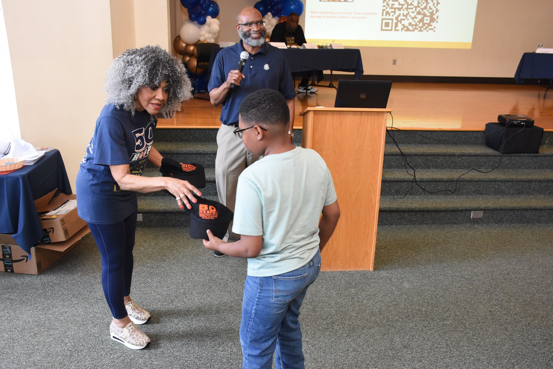 Woman handing a boy a black bag, man speaking at a podium. Event in a room with blue and gold decorations.