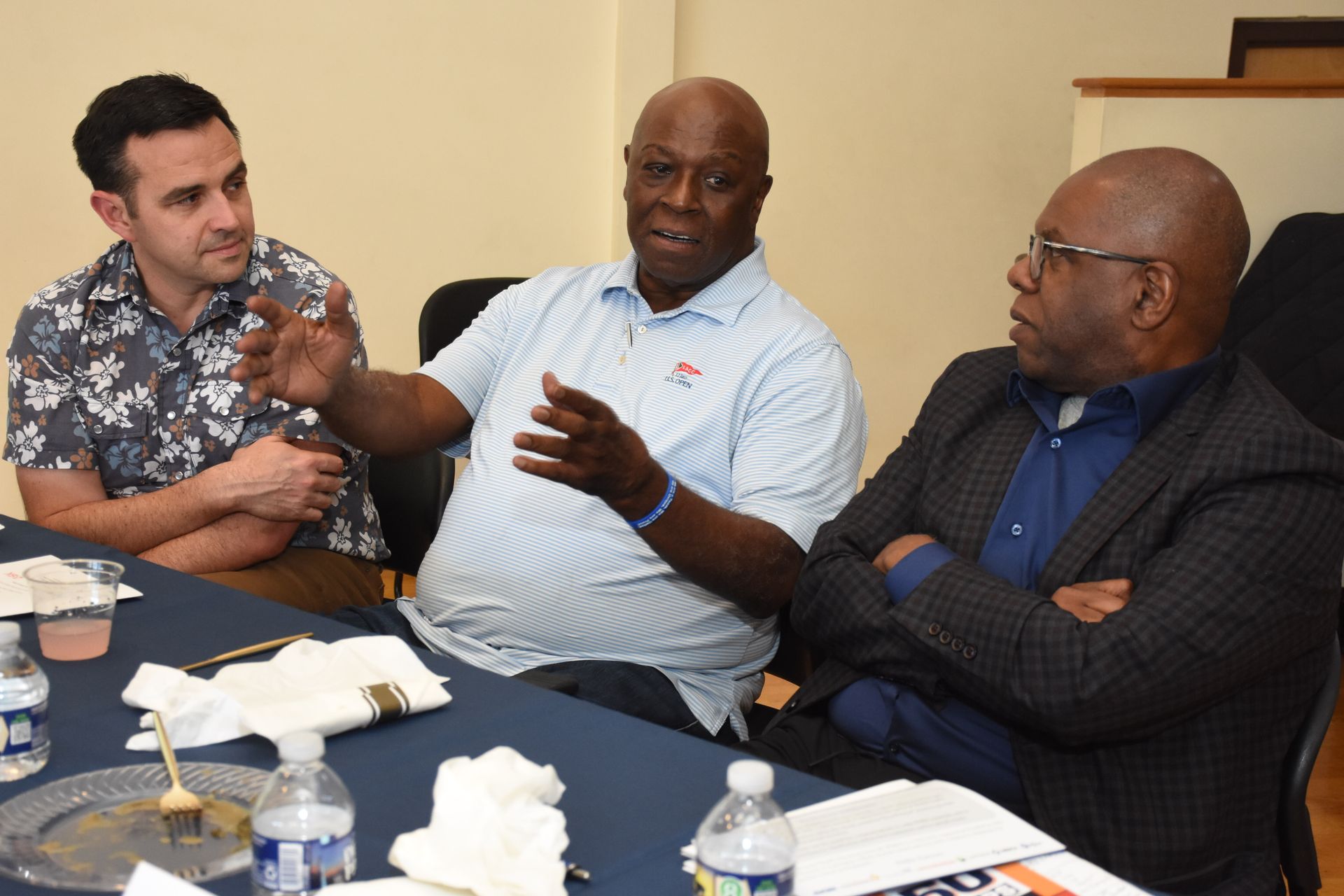 Three men seated at a table. One gestures, speaking. Two others listen. Blue tablecloth, water bottles.