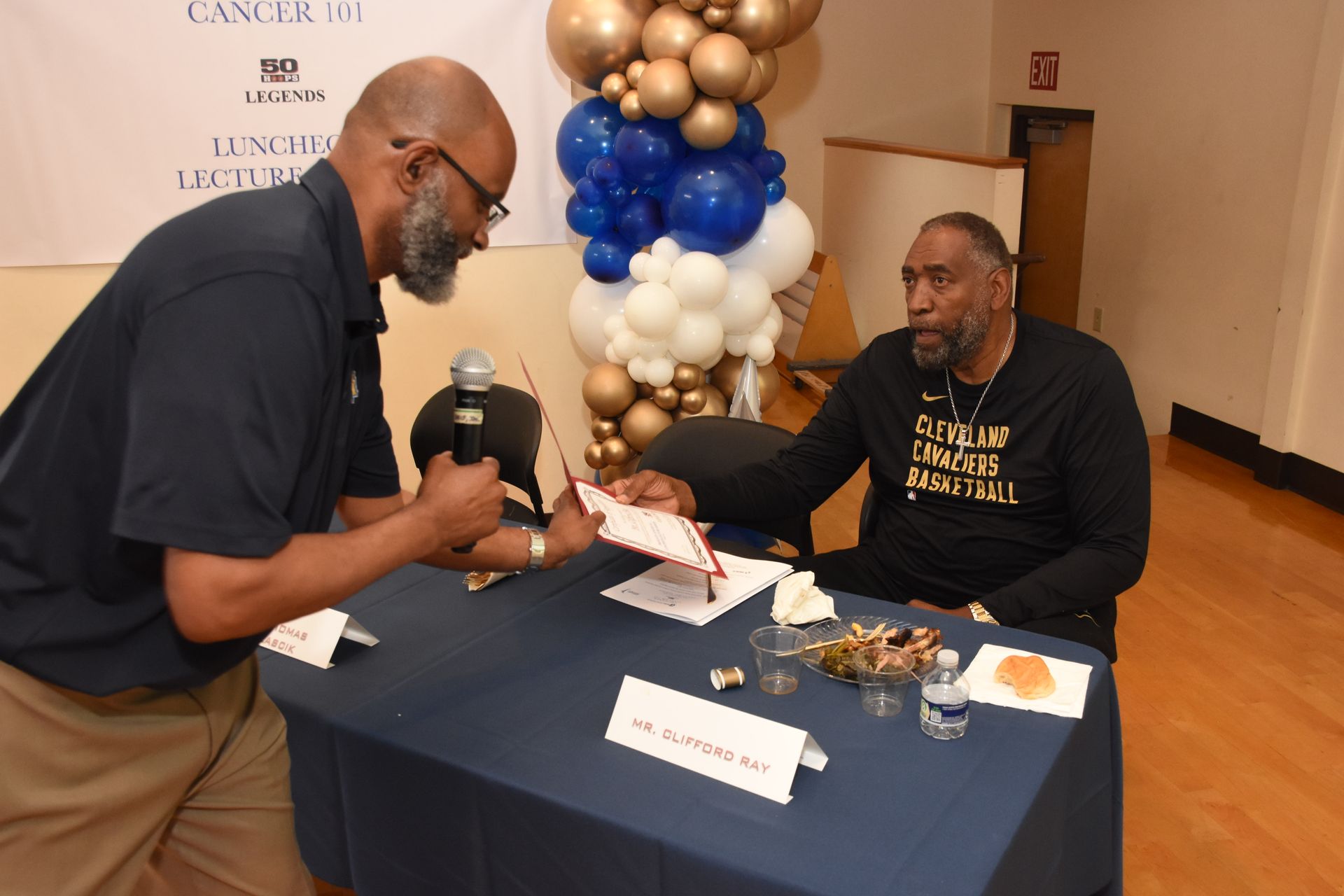 Man signing an autograph, seated at a table. Another man holds a microphone and a paper. Balloons in the background.