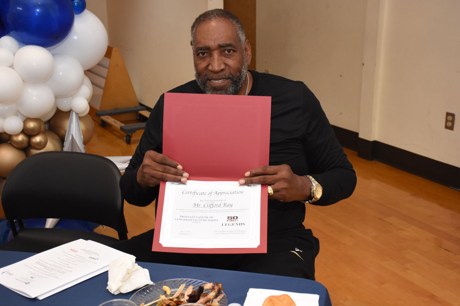 Man in black outfit holds a certificate, smiling. Balloons and food are in the background.