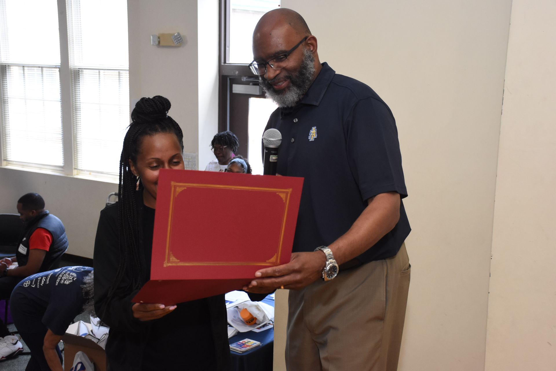 Man presents a certificate to a woman, both smiling. They're indoors.
