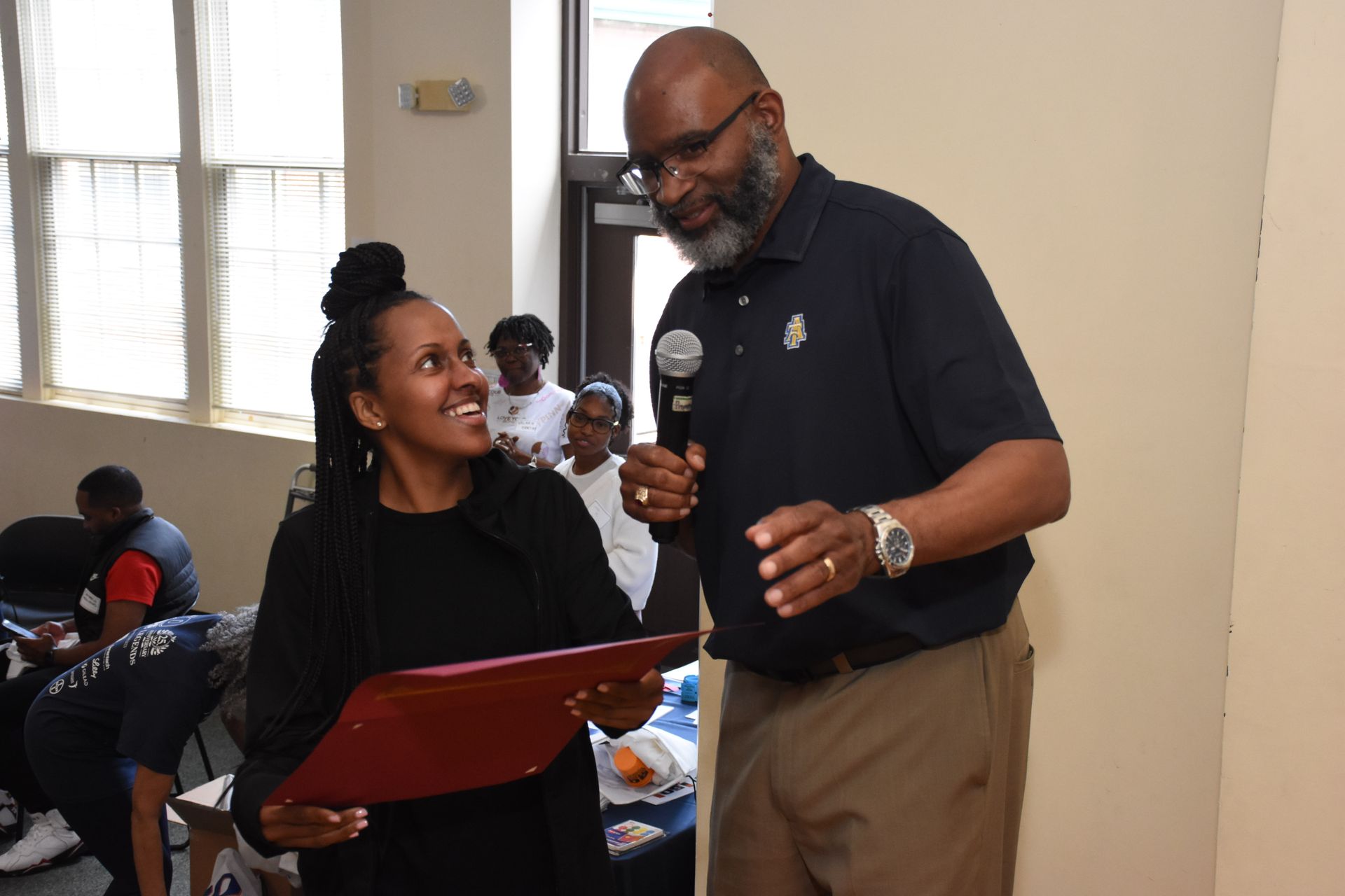 A man hands a woman a certificate. They are smiling. Indoor setting with a crowd in the background.