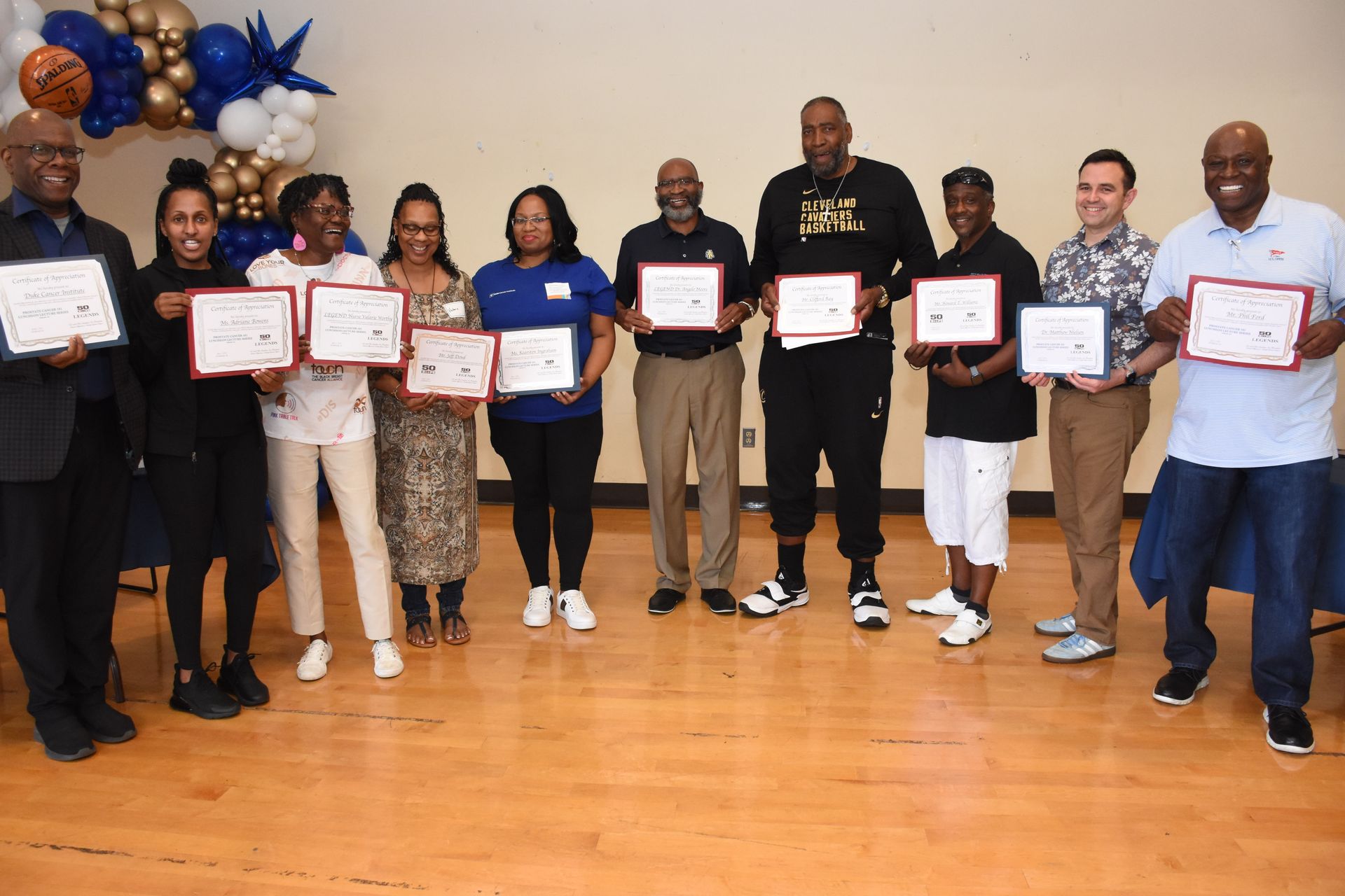 Group of people holding certificates, smiles for a photo. Blue and gold balloons in background.