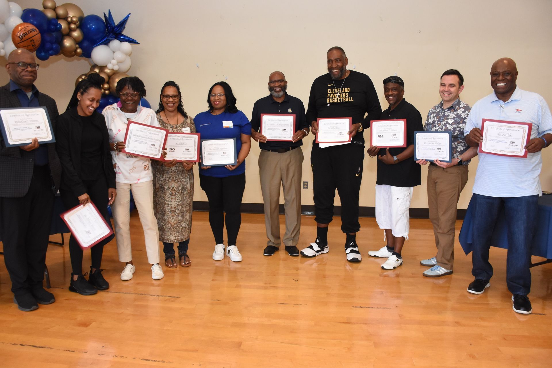 Group of people holding certificates, smiling, indoors with blue and gold decorations.