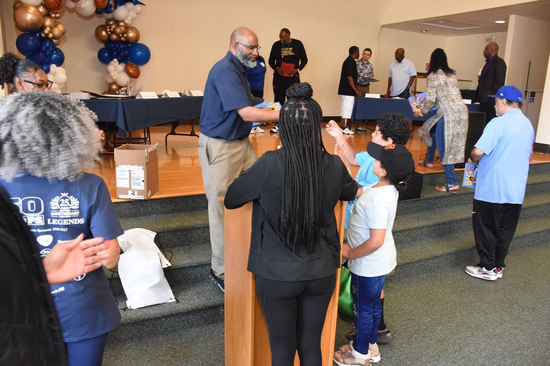 People receiving items from a man at a table, likely a community event. Balloons and tables in a hall.
