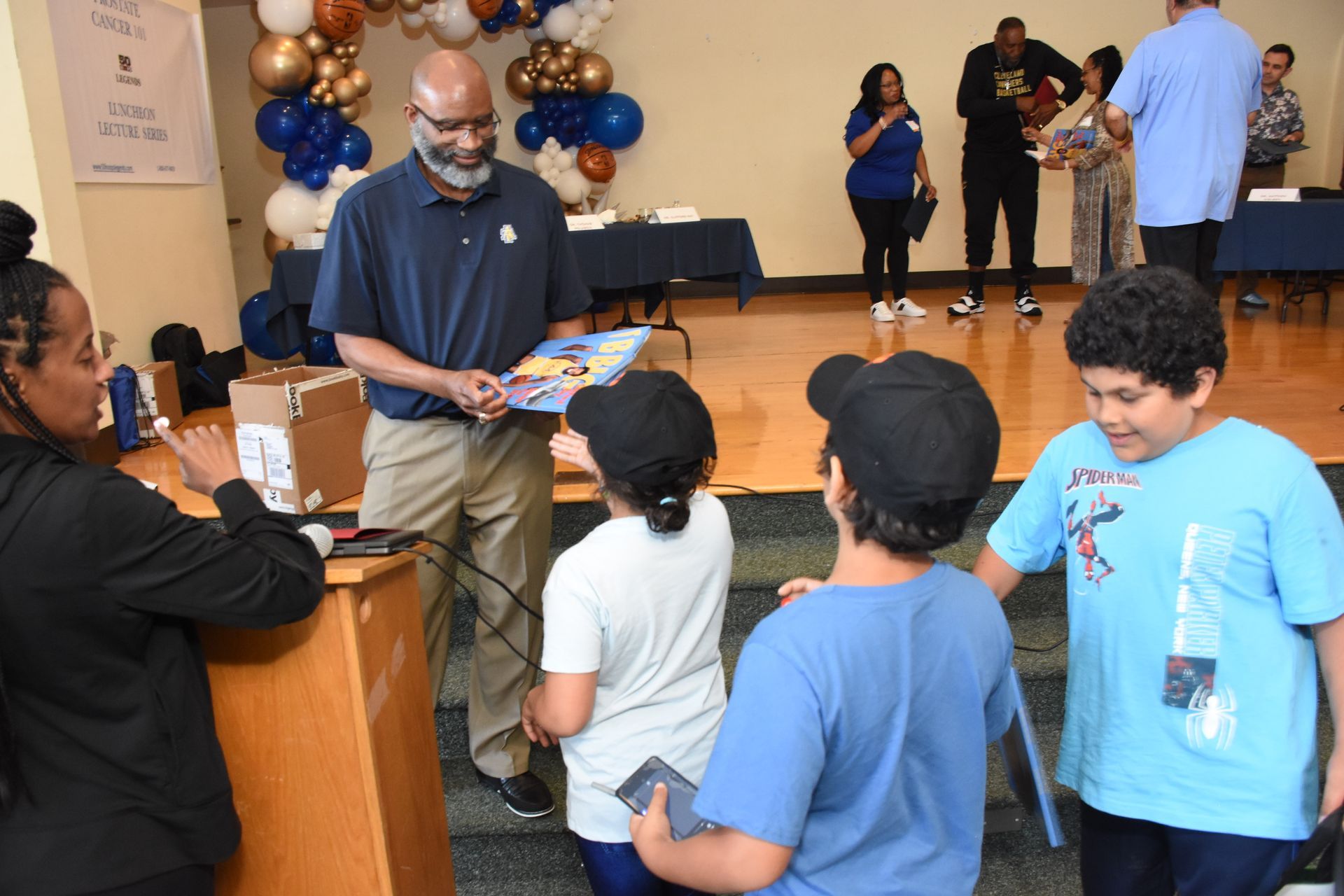 Man giving a book to children at a community event; balloons in background.