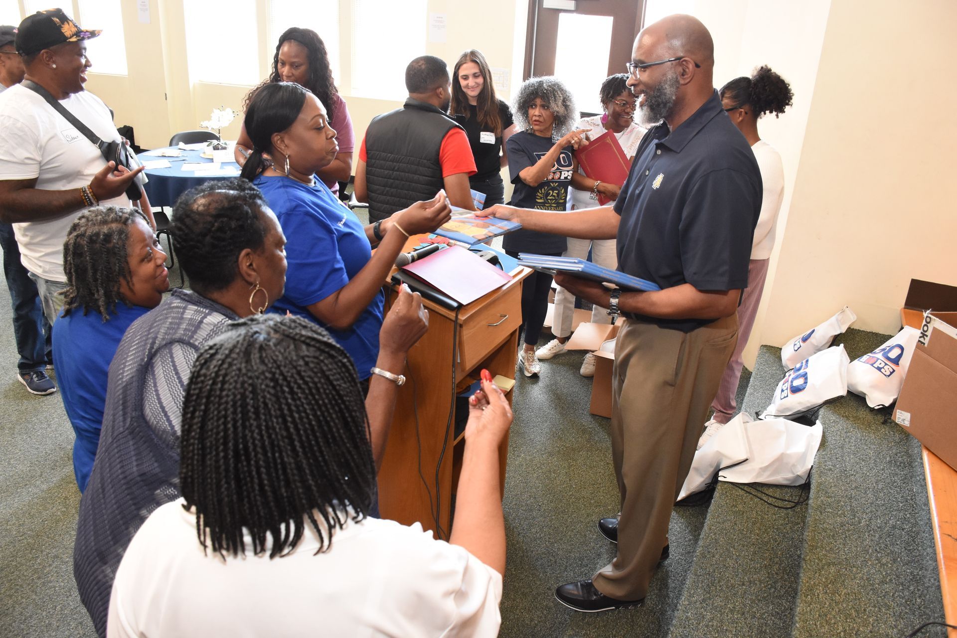 People gathered in a room, a man hands out papers; tables, boxes, and a podium present.