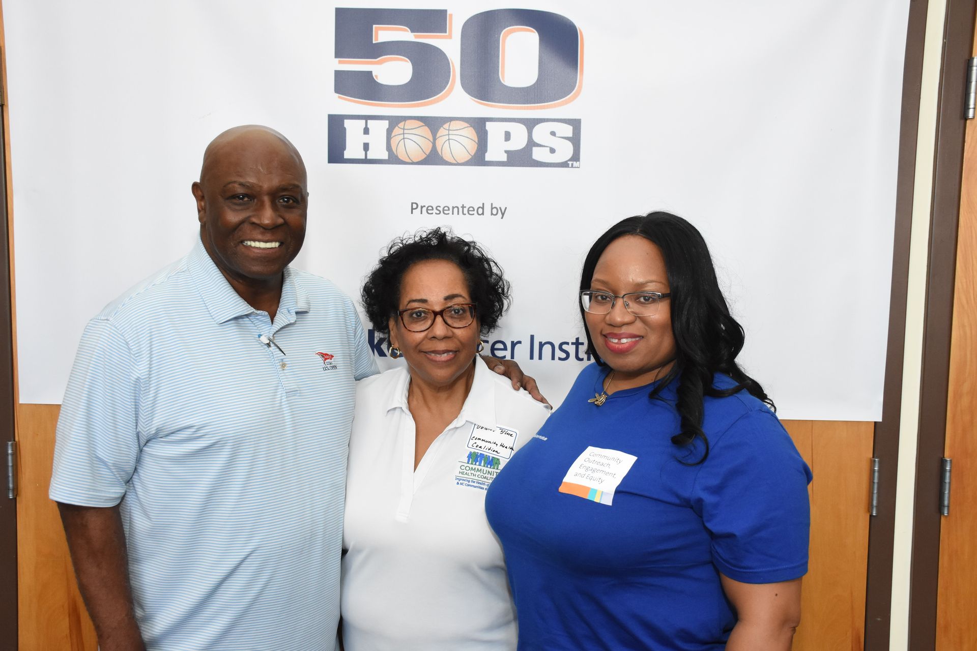 Three people pose in front of a banner. A man in a blue polo smiles beside two women.
