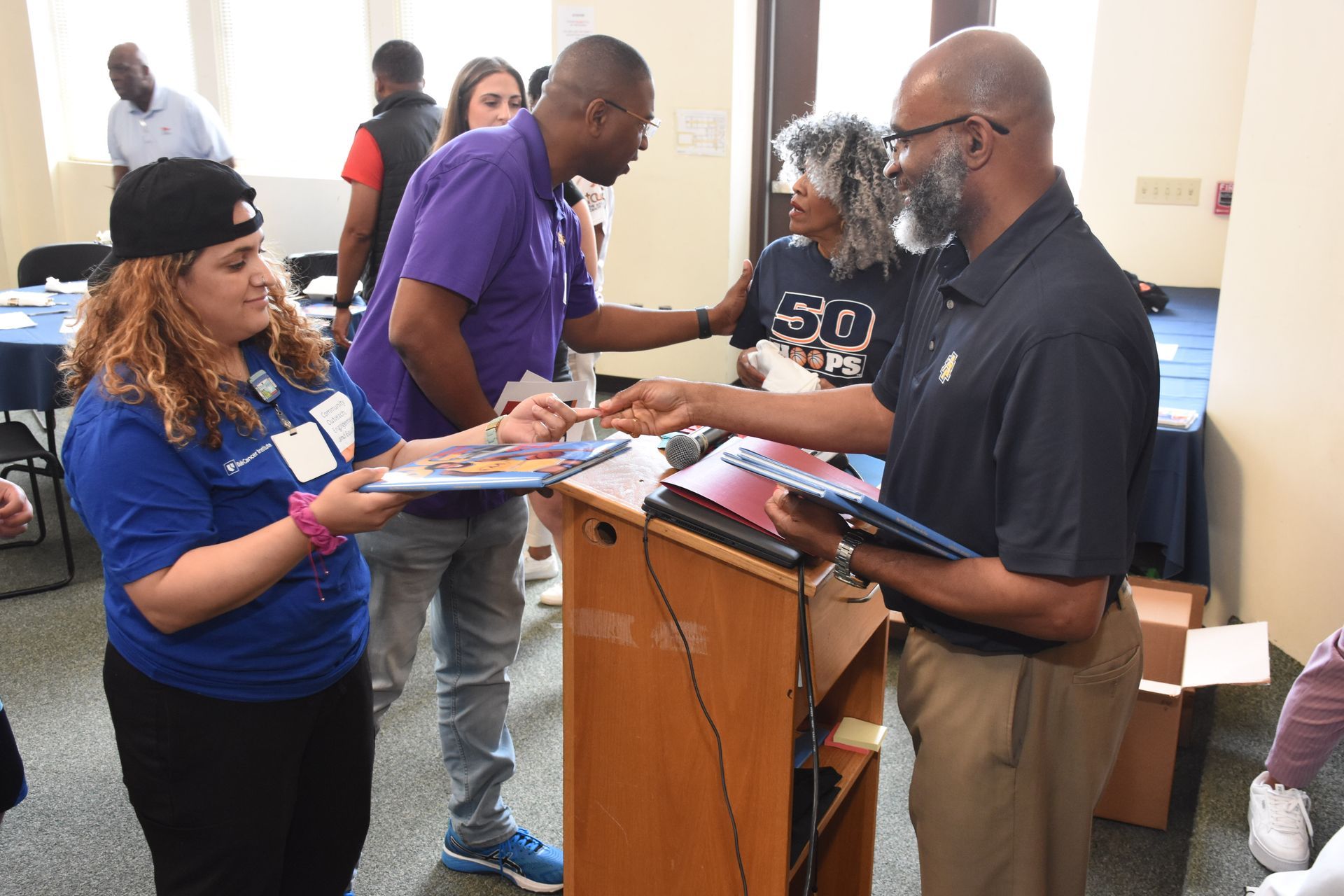 People at an event, exchanging materials near a podium. Several wear blue or purple shirts.