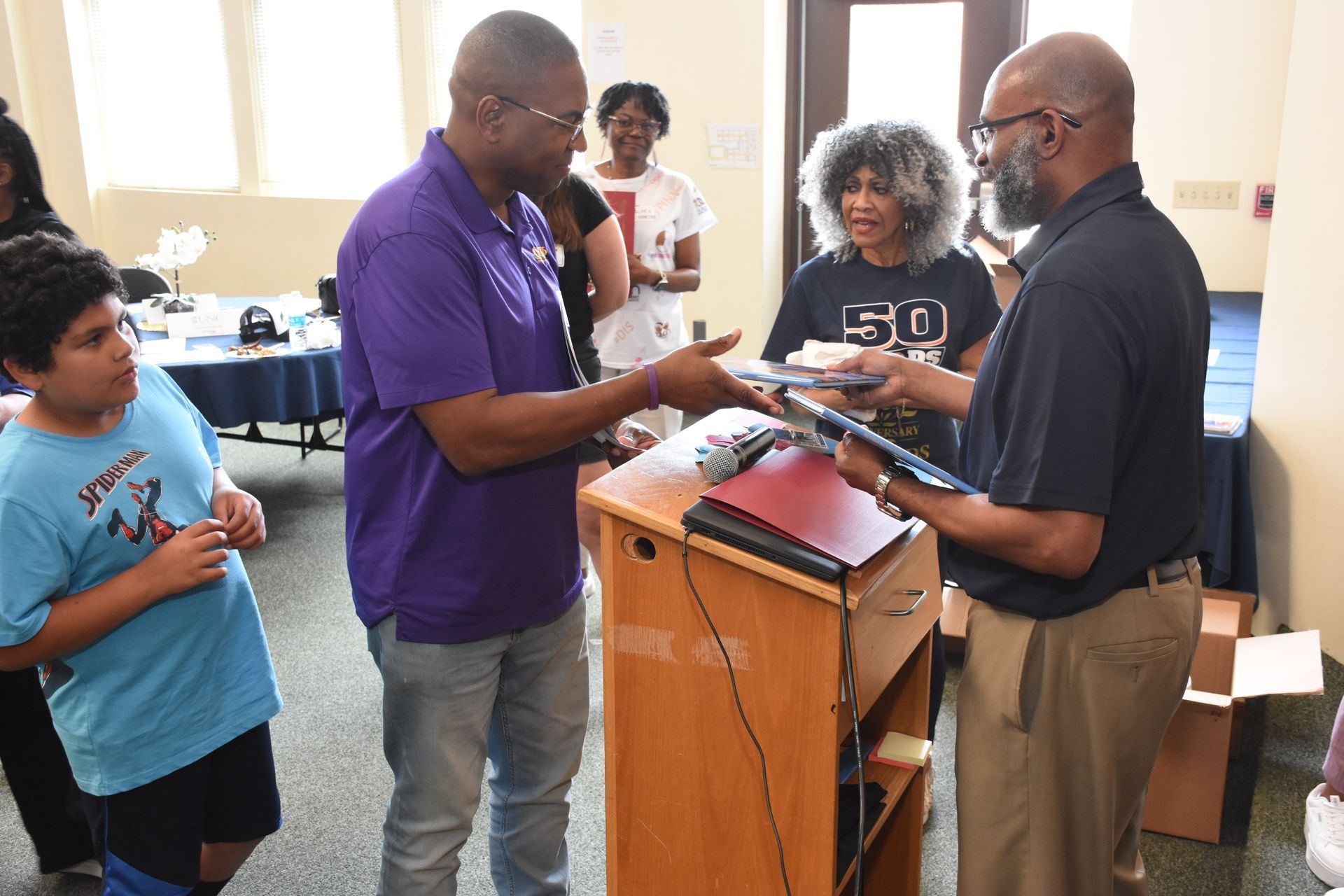 Man in purple shirt hands laptop to another man, a boy watches. Setting indoors, other people present.