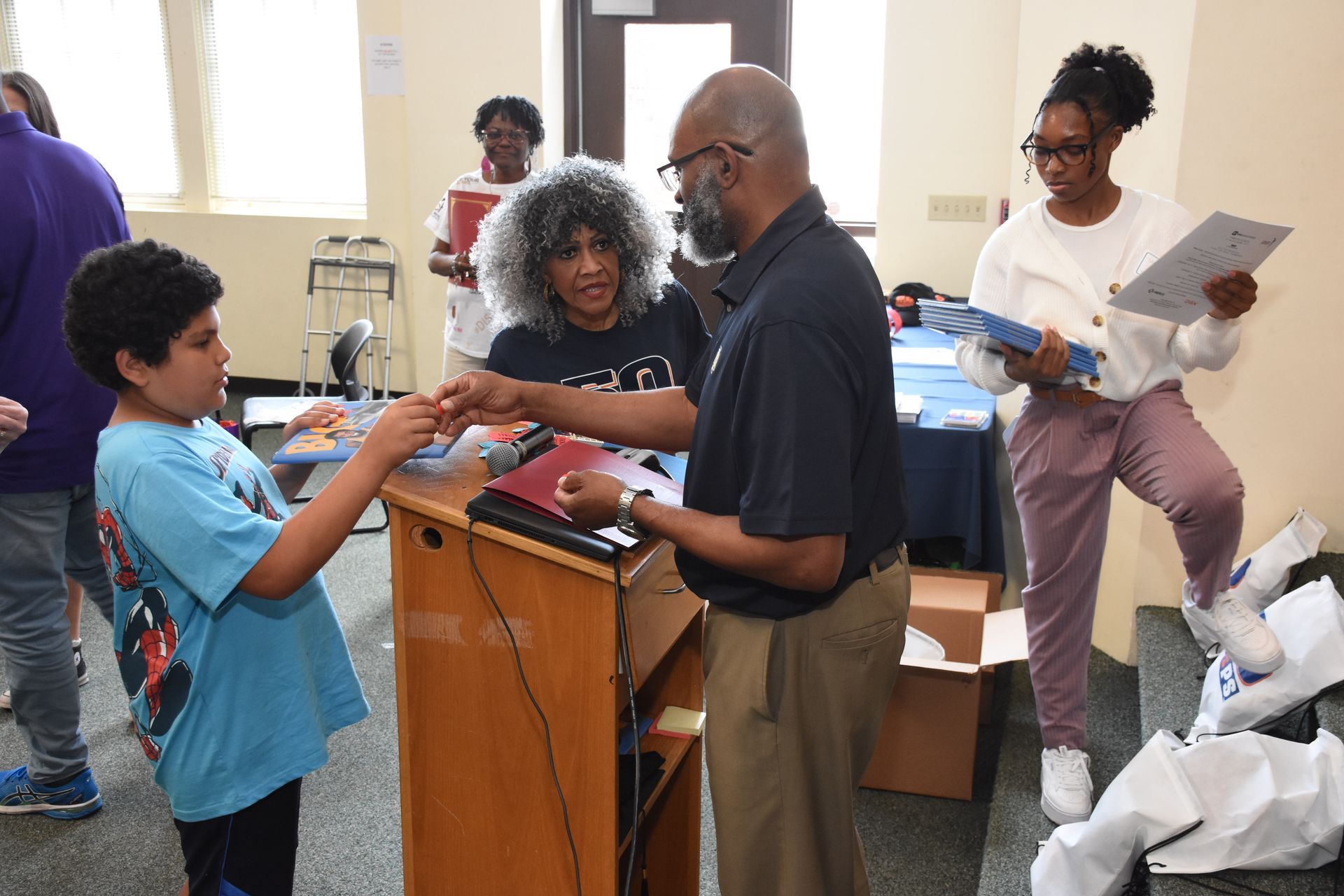 Man handing a young person a book at a podium, others present, interior room.