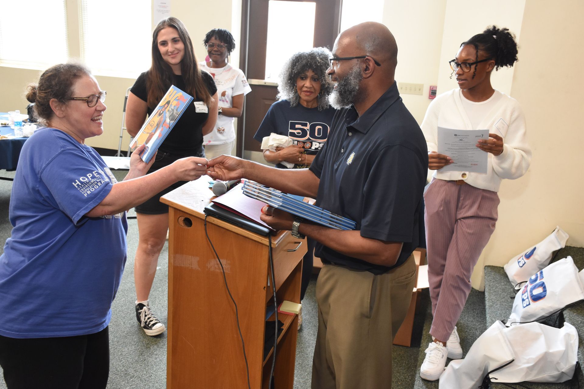 Woman in blue shirt hands a book to a man behind a podium, other people watch. Indoors, bright light.