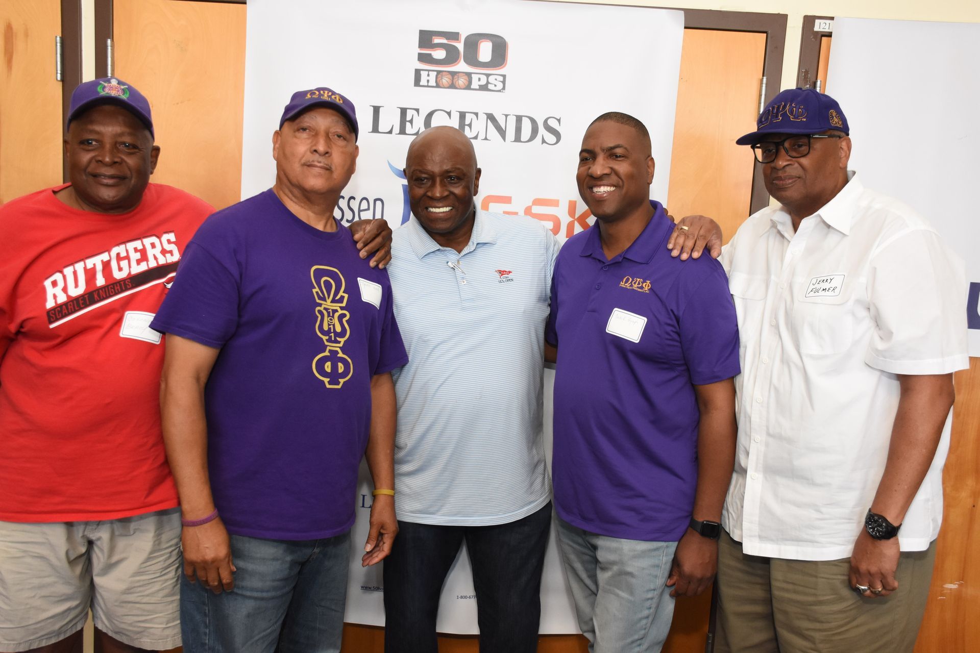 Five men pose together indoors; some wear purple shirts. A banner reads 