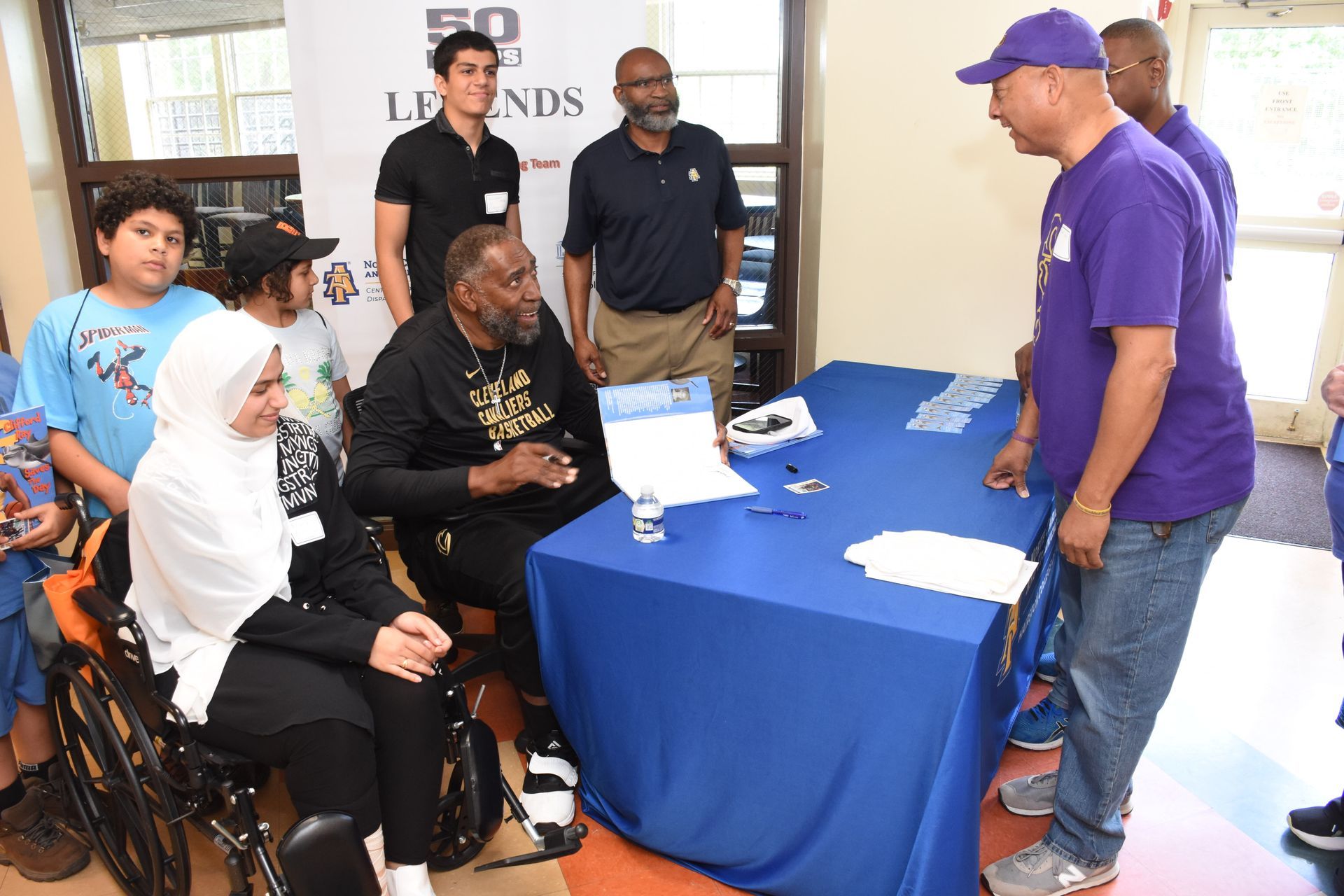 People at a table. A man in a wheelchair, surrounded by others, interacts with a man in purple.