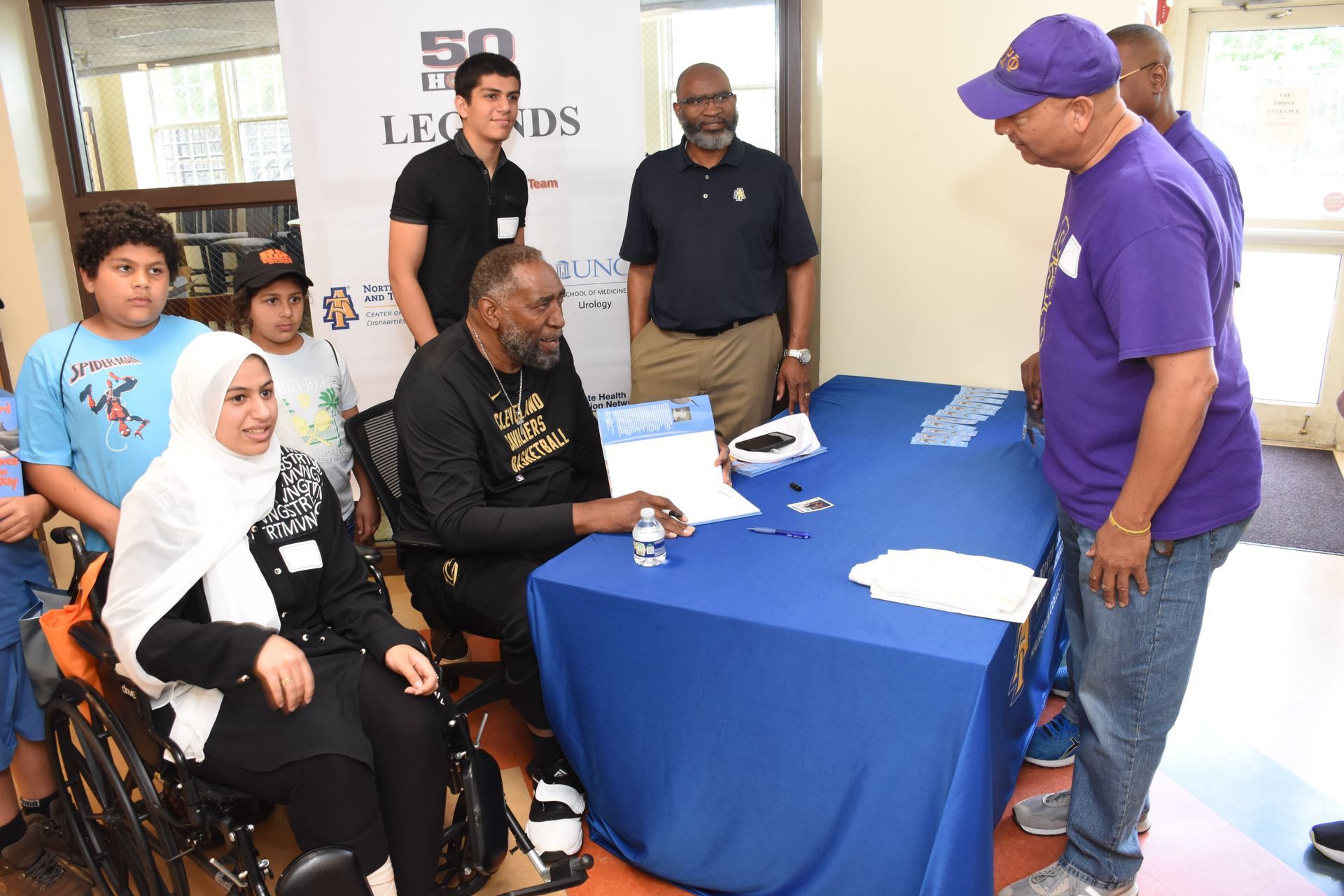 People at a table; one signing documents. Others watch, some in purple, inside a building.