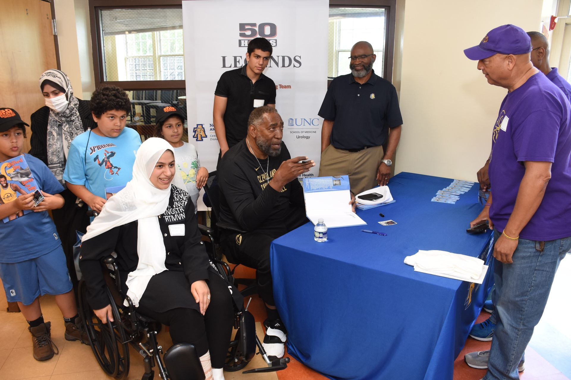 People gather around a table with a man signing papers. Children and adults observe.