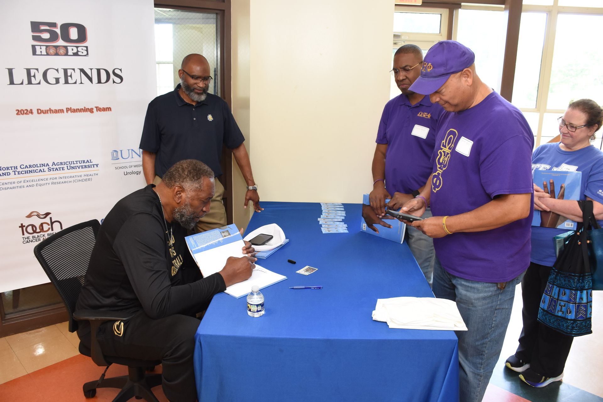 Man signing at a table, two others look on; event banner in background.