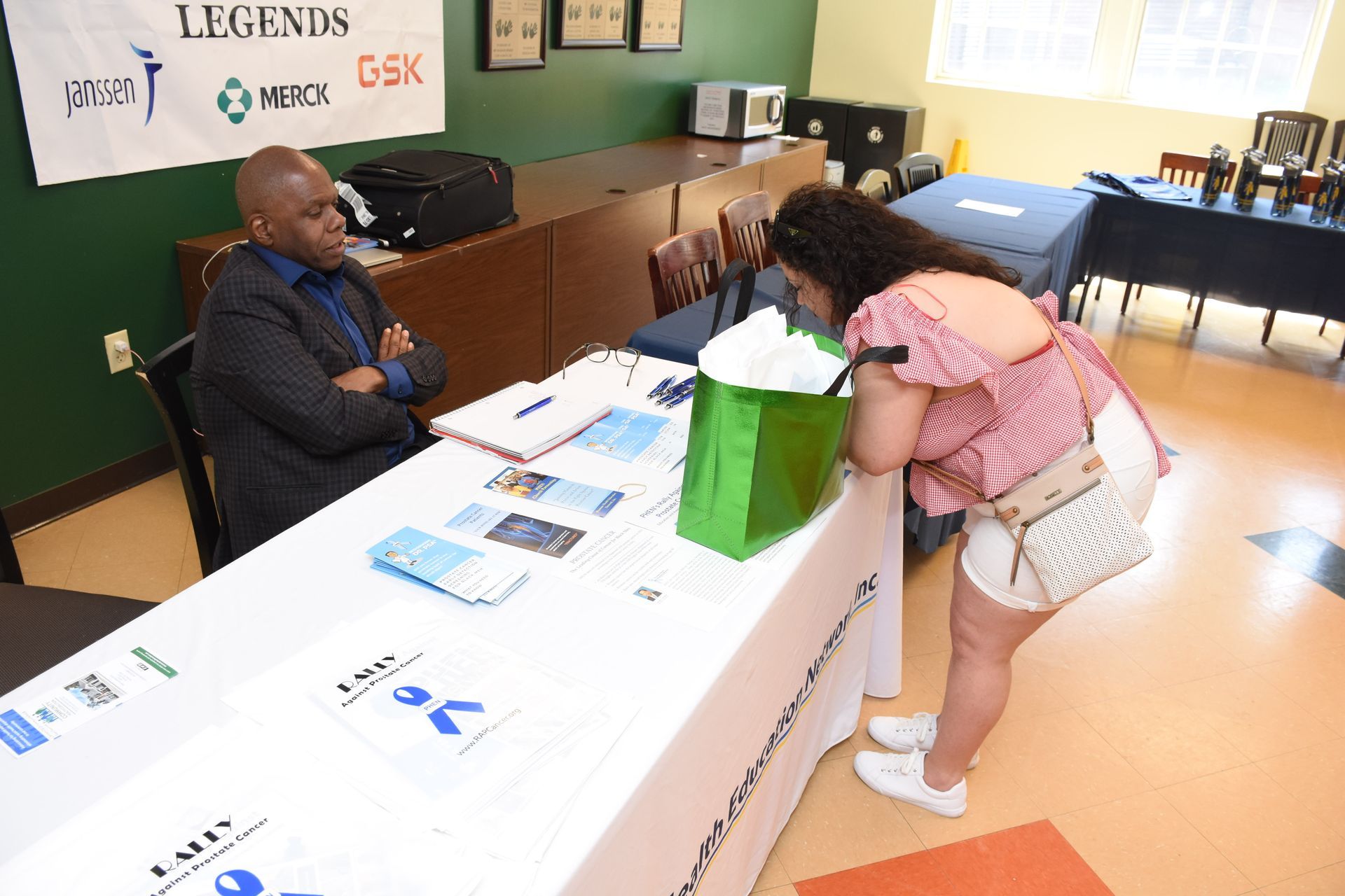A person leans over a table with printed materials while another person sits and looks on. Green gift bag on table.