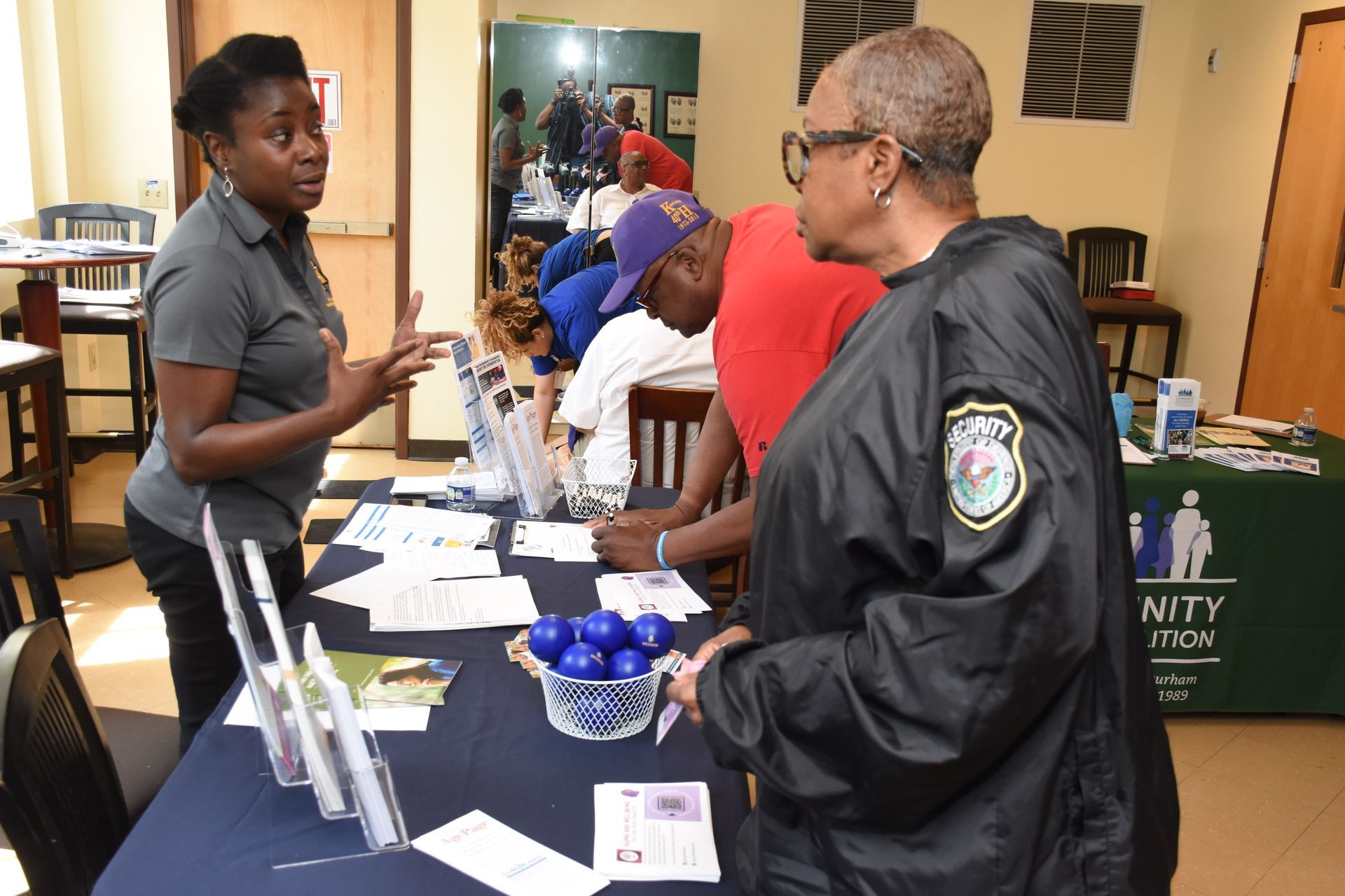Woman at table with brochures talking to a person, others in the background.
