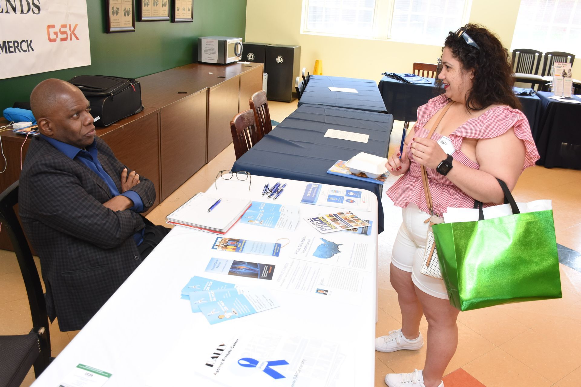 Man seated at a table with a woman standing, looking at materials. Blue and white documents are on the table.