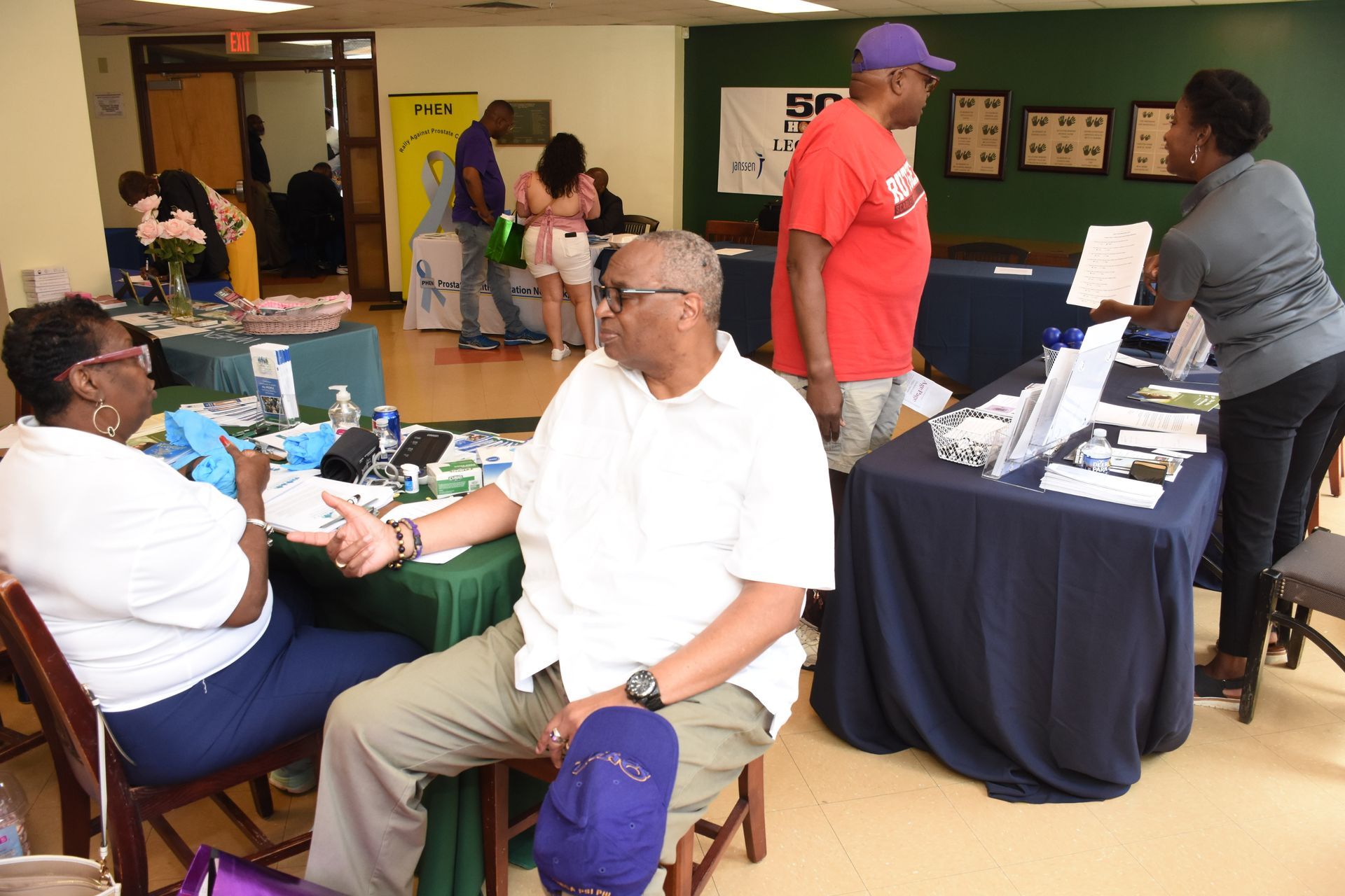 People at health fair, checking vitals, looking at displays. Tables with information in room.