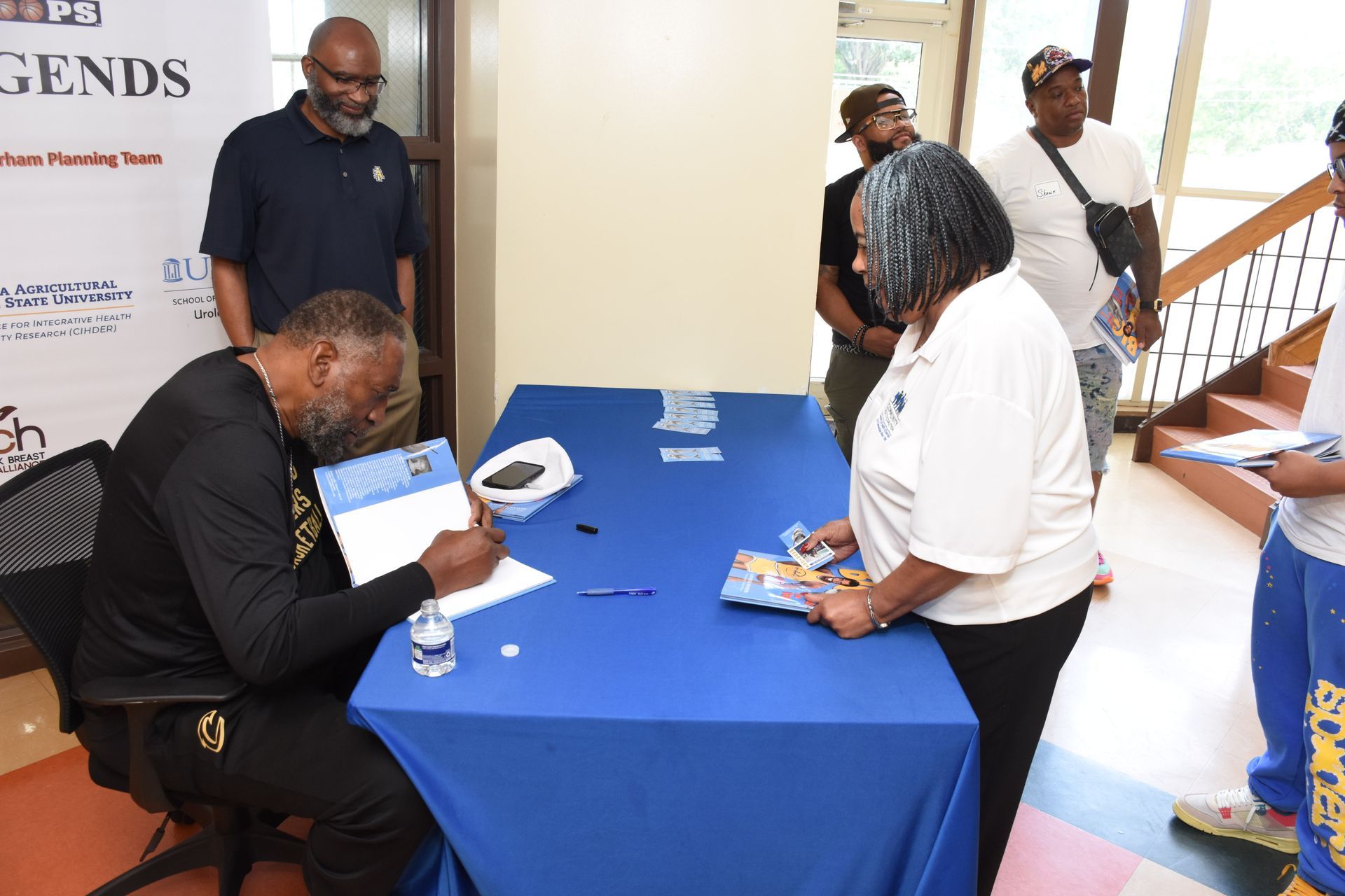 Person signing autographs at a table with a line of people waiting. Blue tablecloth, water bottle, and pens are visible.