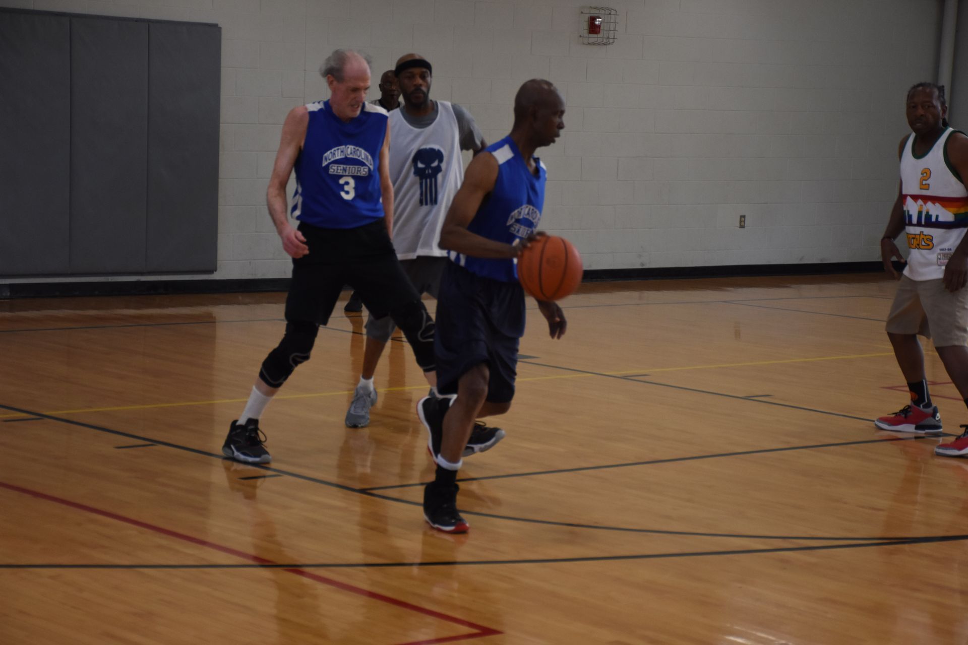 Basketball players on a court. One dribbles, others defend. Wooden floor, neutral background.